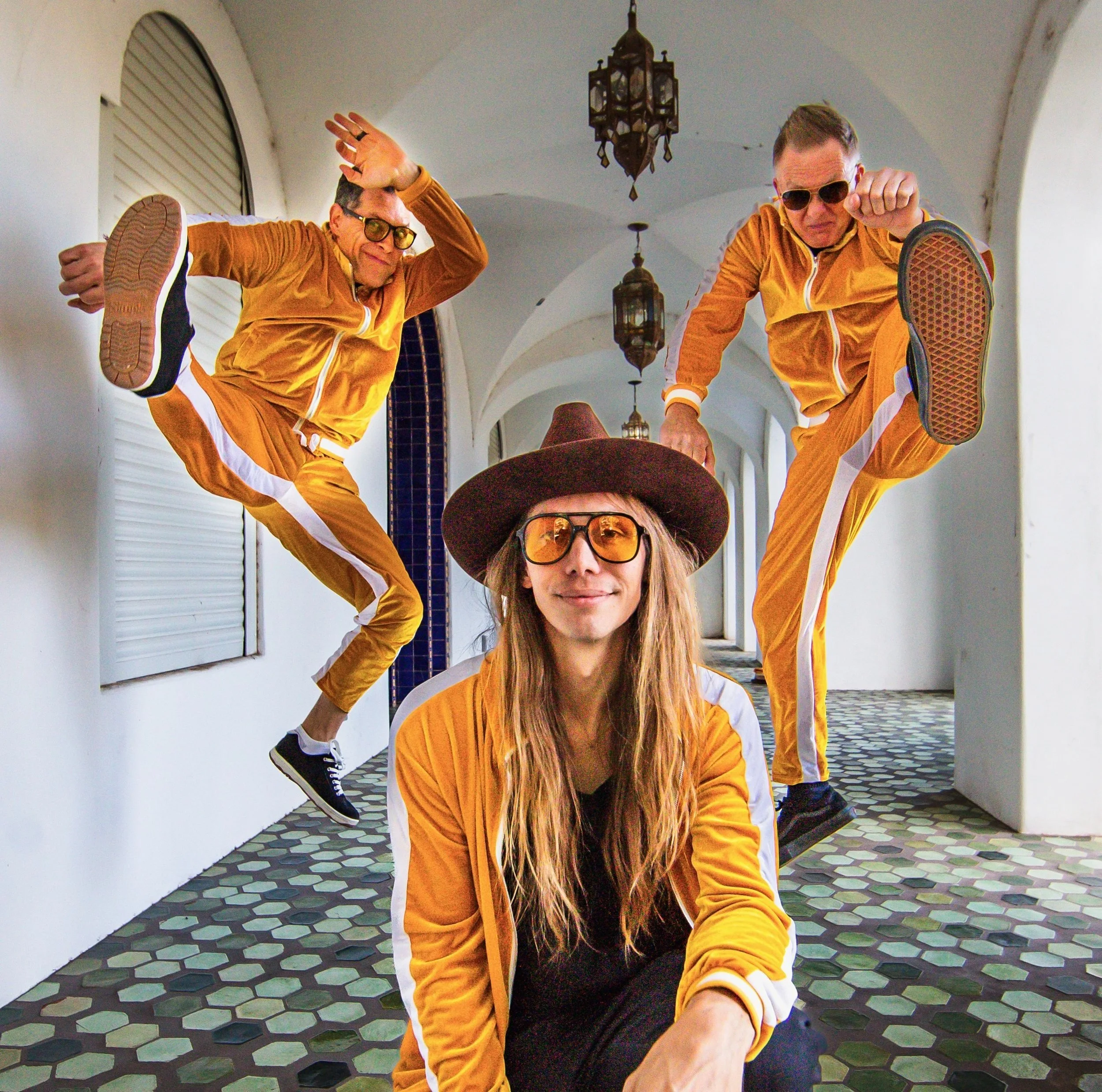 Three people in matching orange tracksuits and sunglasses, two jumping in the air with one person sitting on the ground, in a hallway with white arches and hanging lanterns.