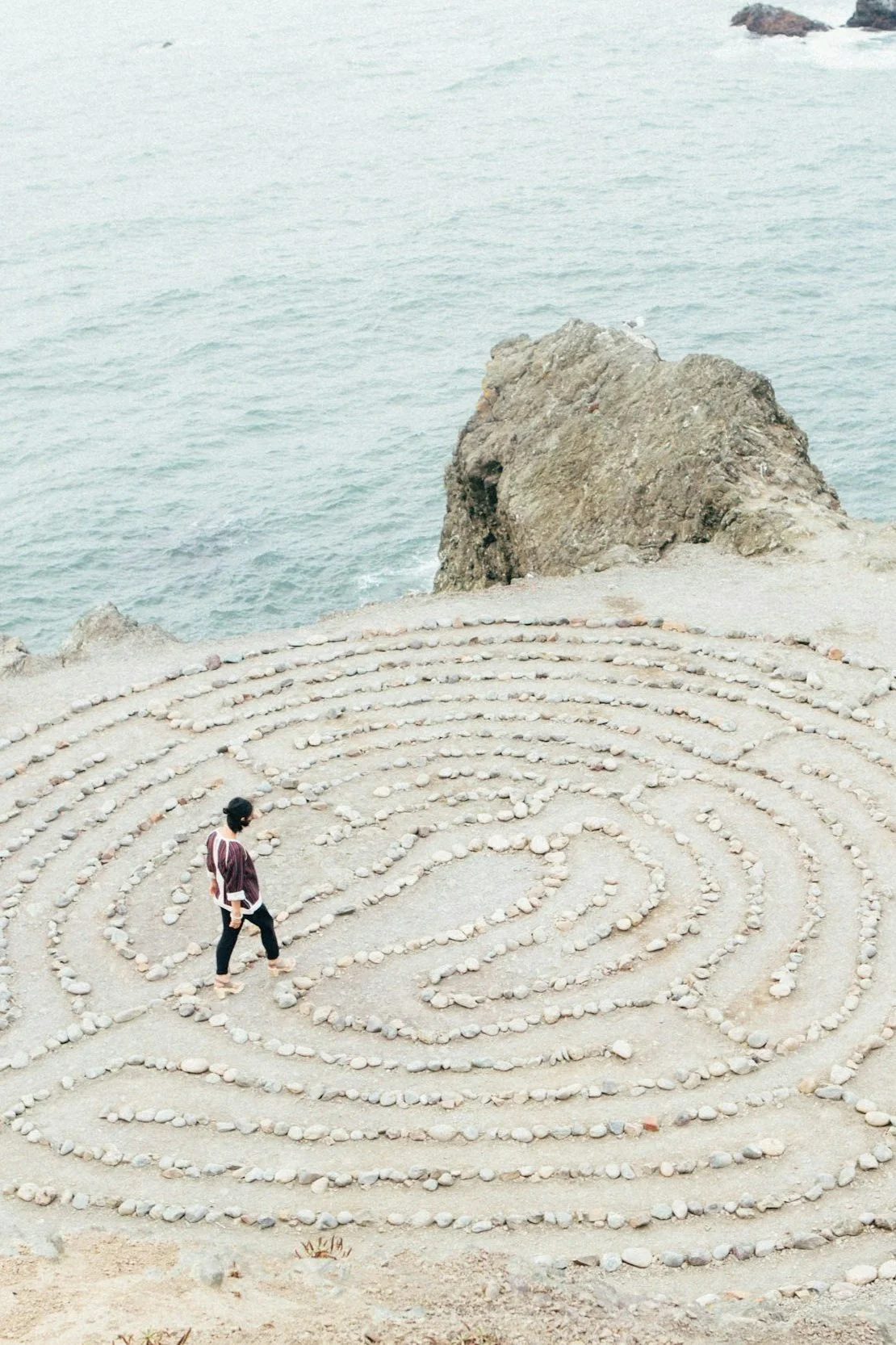 A woman walking through a spiral stone labyrinth on a sandy beach with ocean and rocks in the background.
