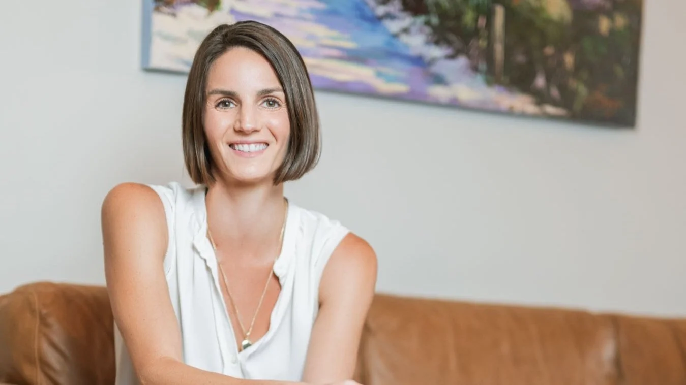 A woman with short brown hair, smiling, sitting on a brown leather couch in a room with a colorful landscape painting on the wall behind her.
