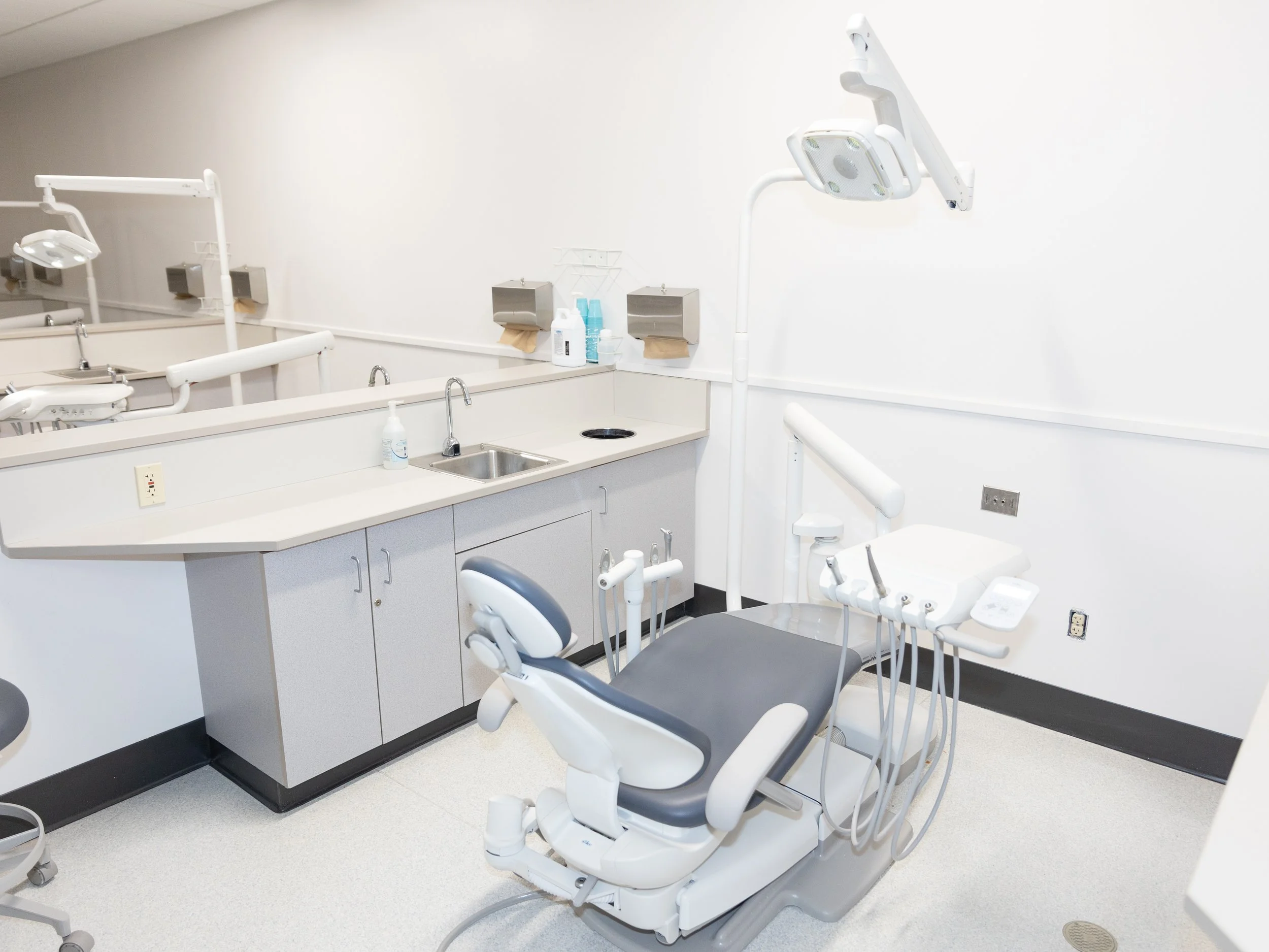 Dental exam room with dental chair, overhead light, sink, cabinets, and dental equipment in a clinical setting.