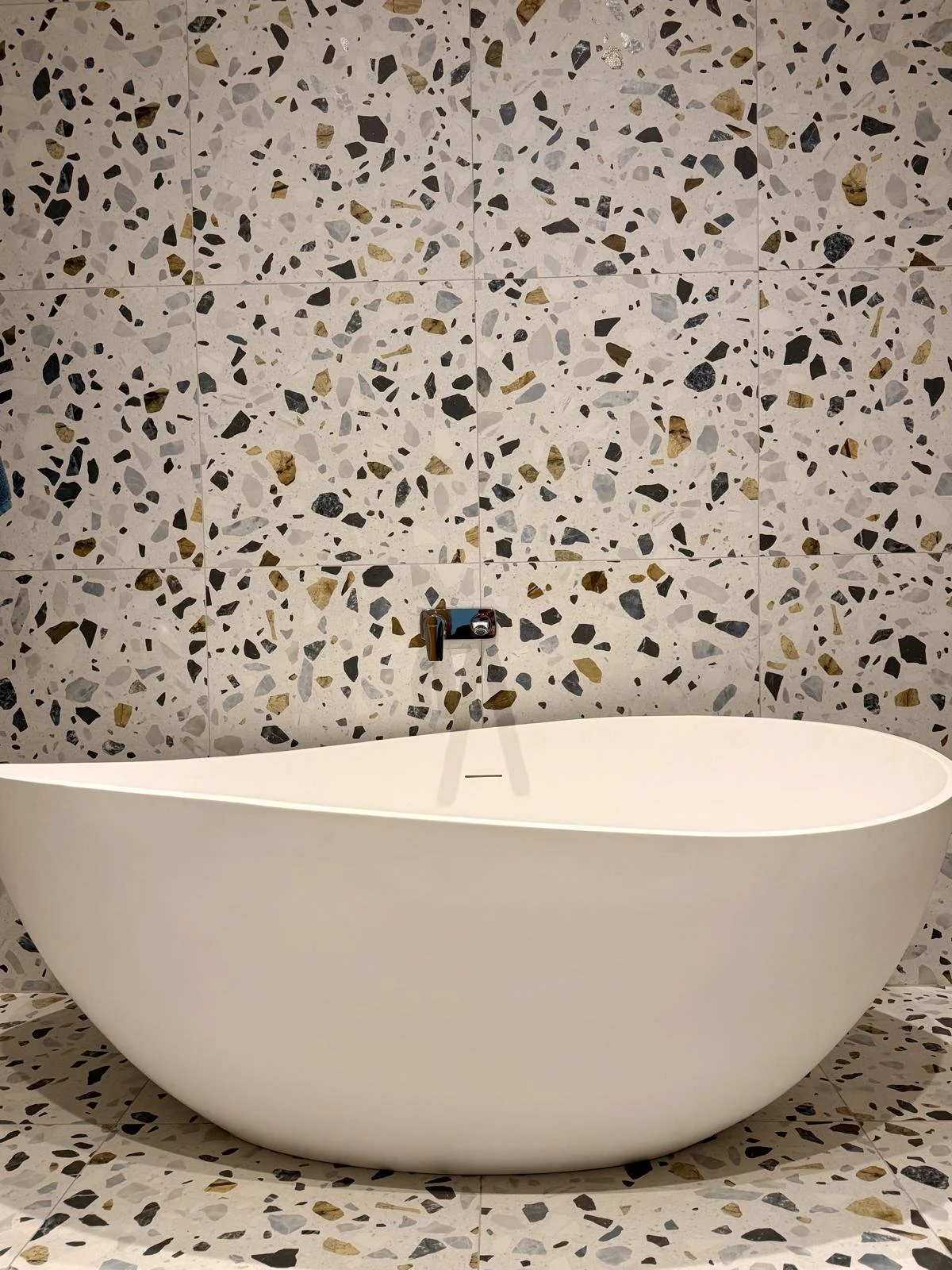 Modern white freestanding bathtub with oval shape, set against a terrazzo wall and floor tiles.