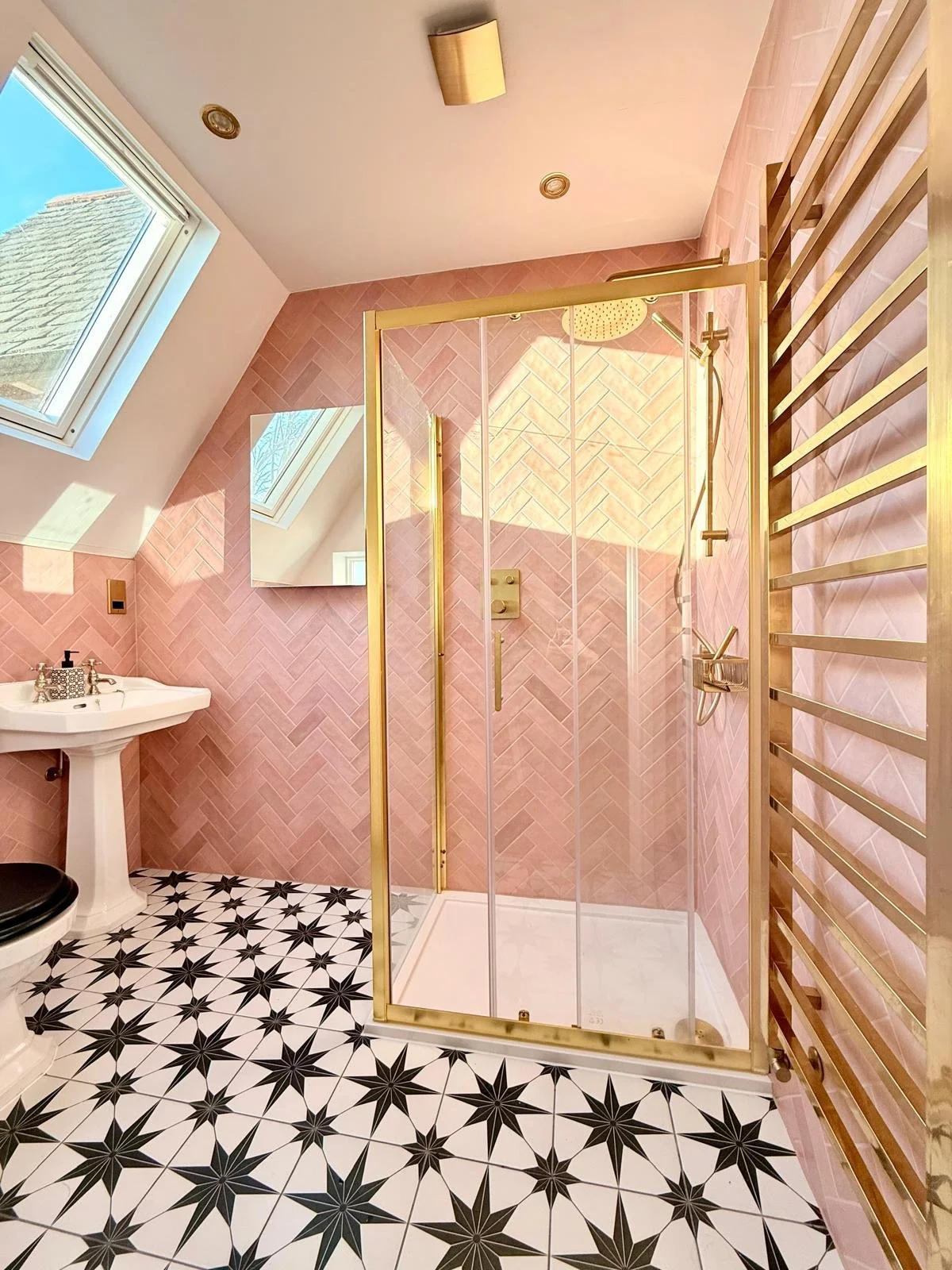 Bathroom with pink tiled walls, a glass shower enclosure with gold trim, a black toilet, a small white pedestal sink, a wall mirror, a skylight window, and a heated towel rack.