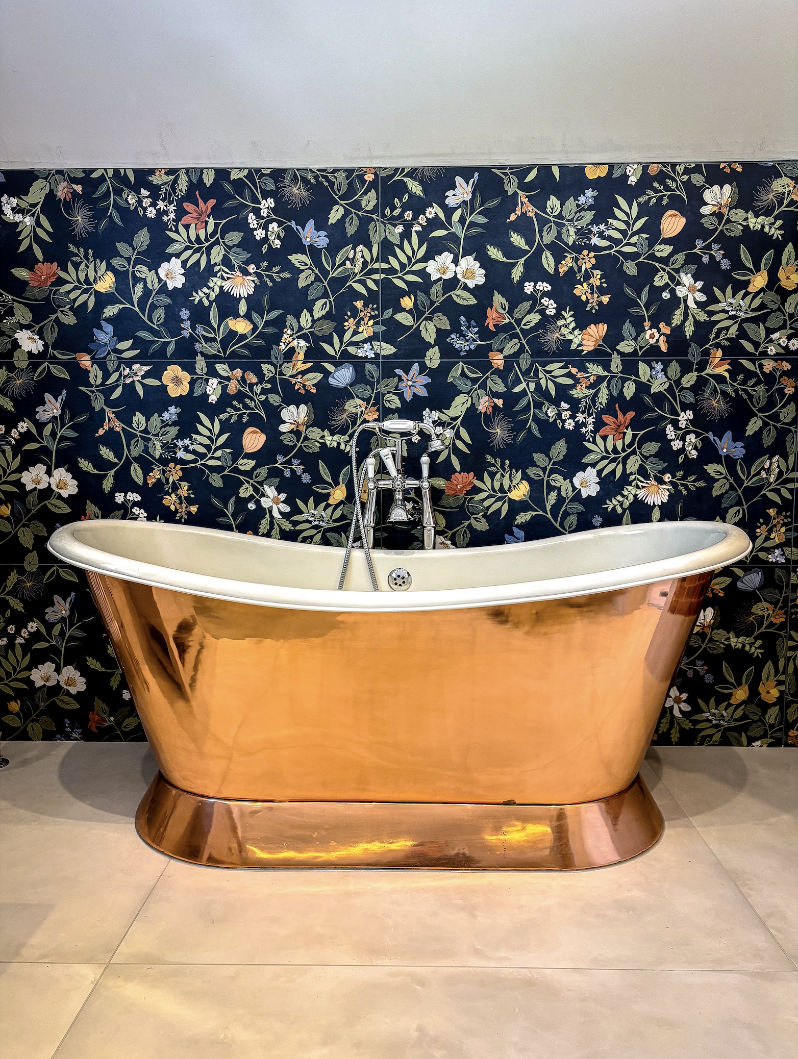 A copper clawfoot bathtub with a vintage style, placed in front of a dark floral wallpaper featuring various flowers and leaves.