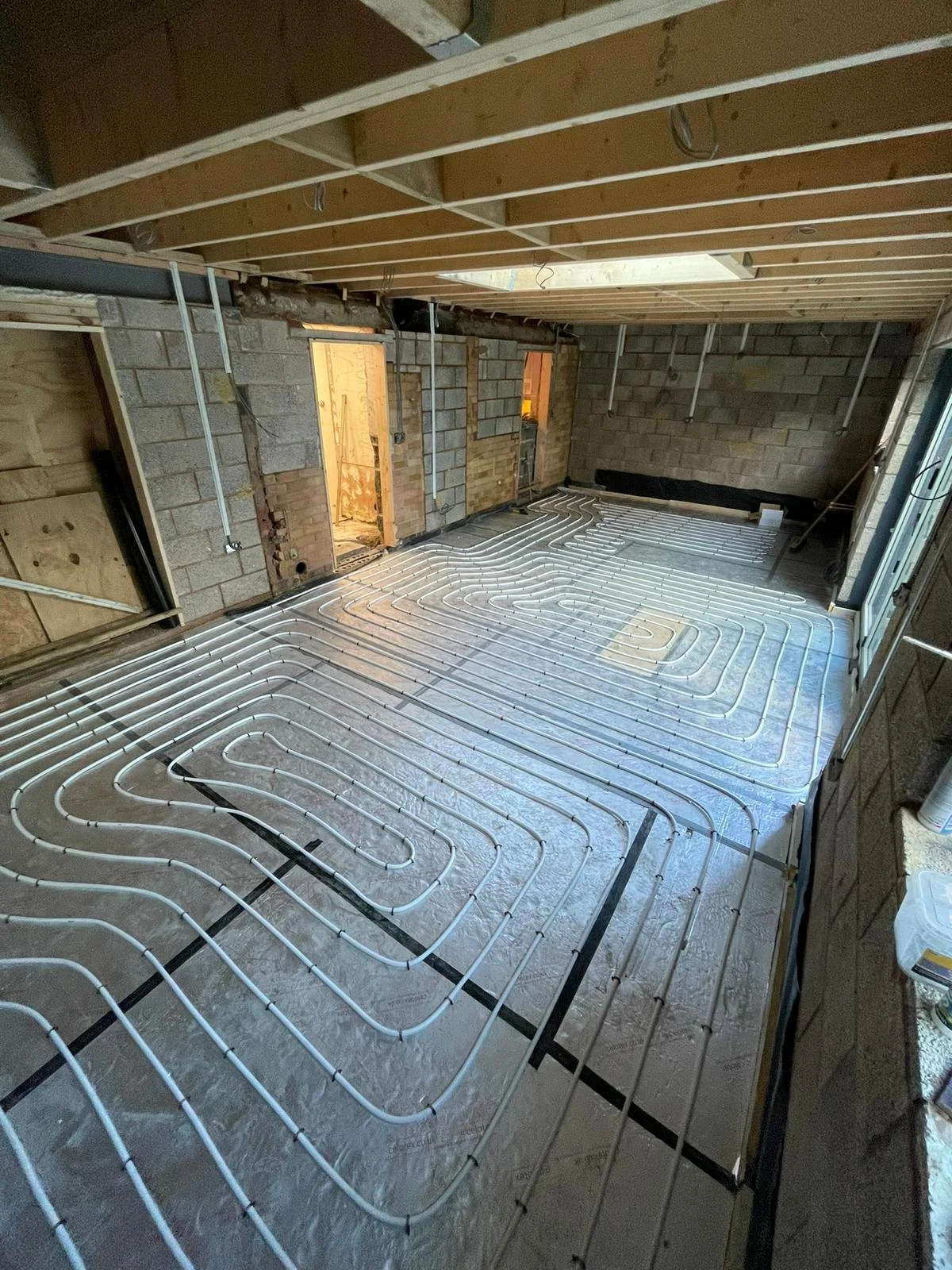 Room under construction with electrical or plumbing underfloor heating pipes installed on the floor, unfinished walls, and exposed ceiling framework.