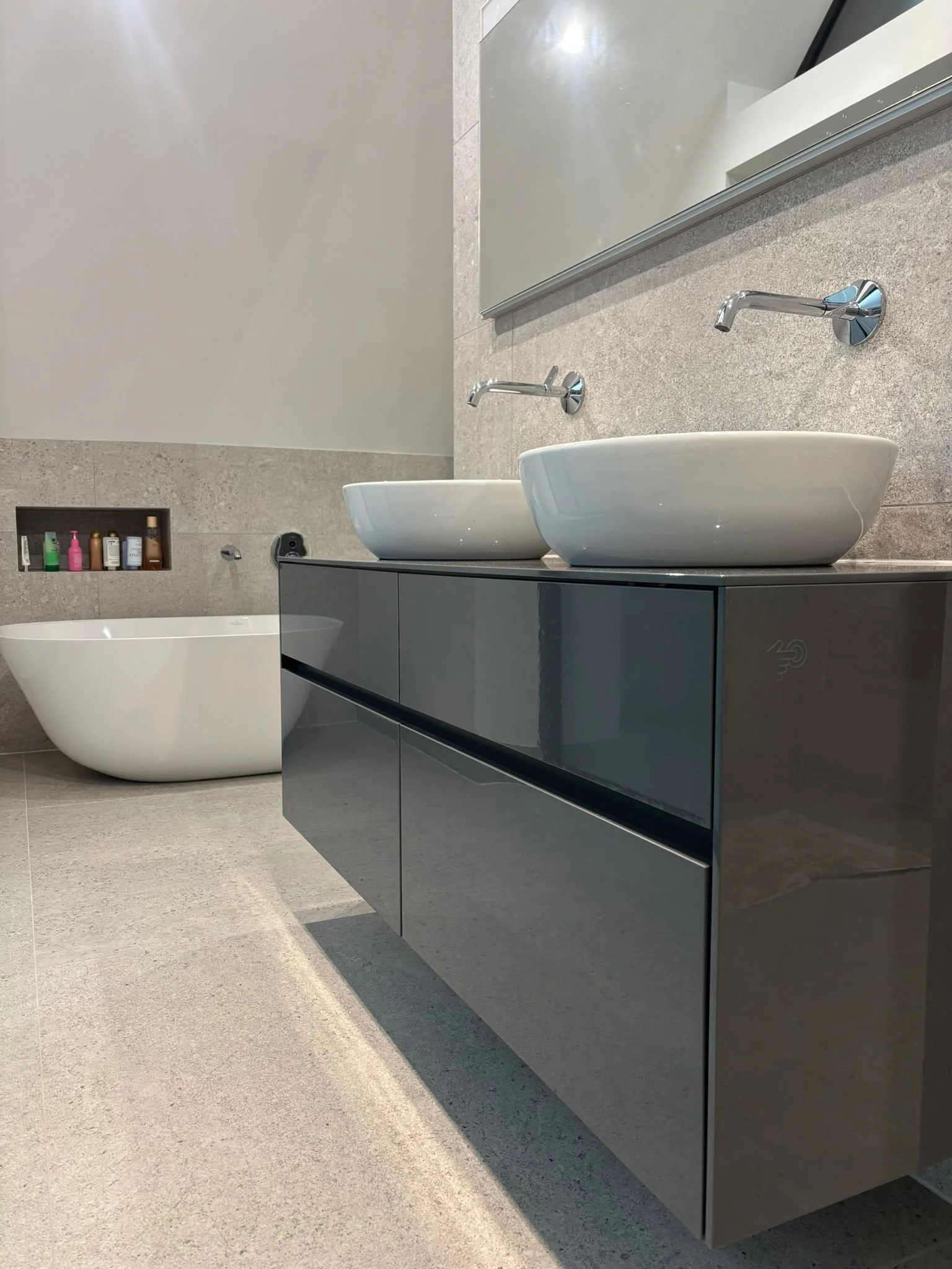 Modern bathroom with double vessel sinks on a glossy black cabinet, wall-mounted faucets, large mirror, and a built-in niche with toiletries in the background.