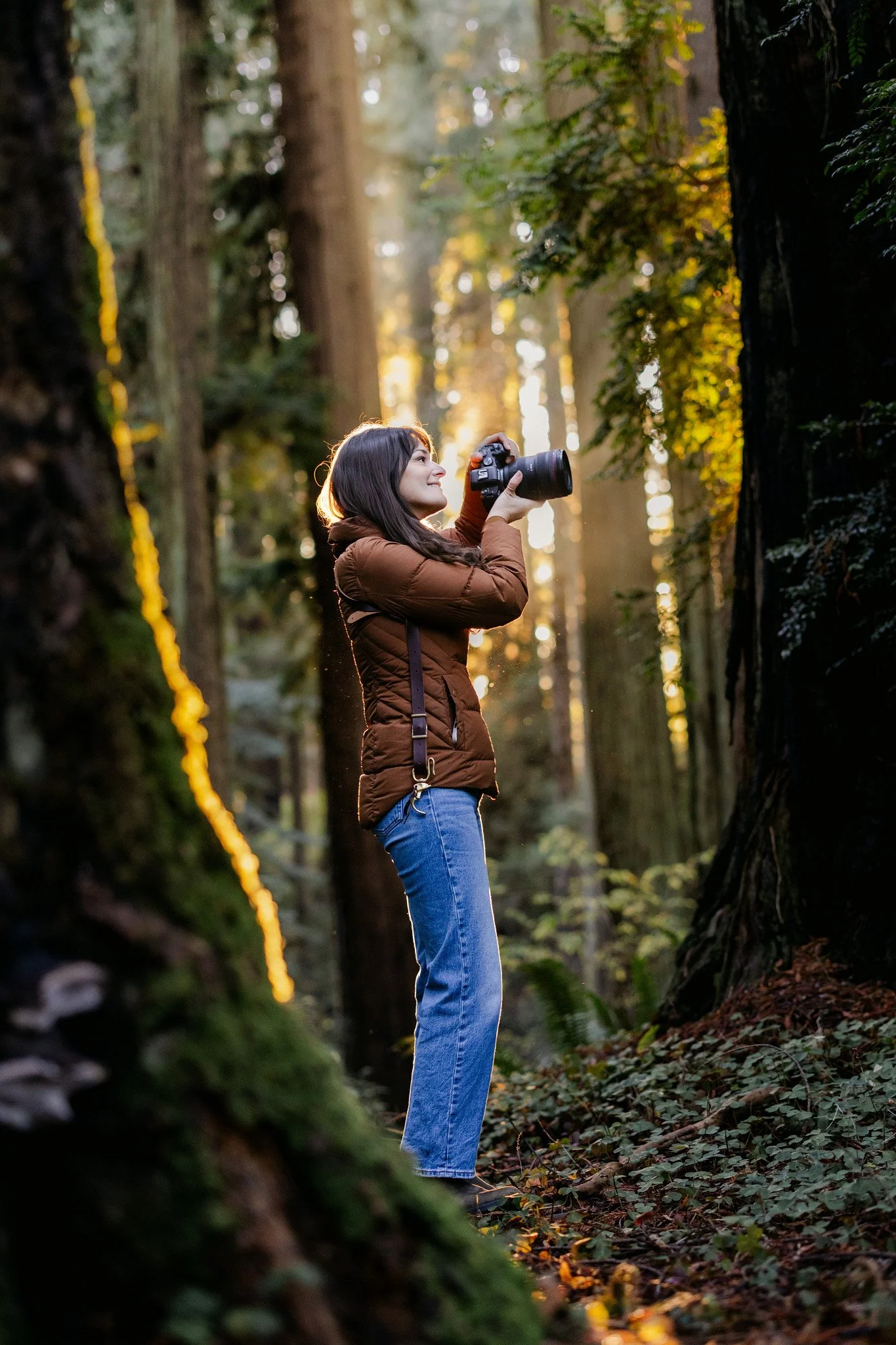 A woman in a brown jacket and blue jeans holding a camera in a forest at sunset.