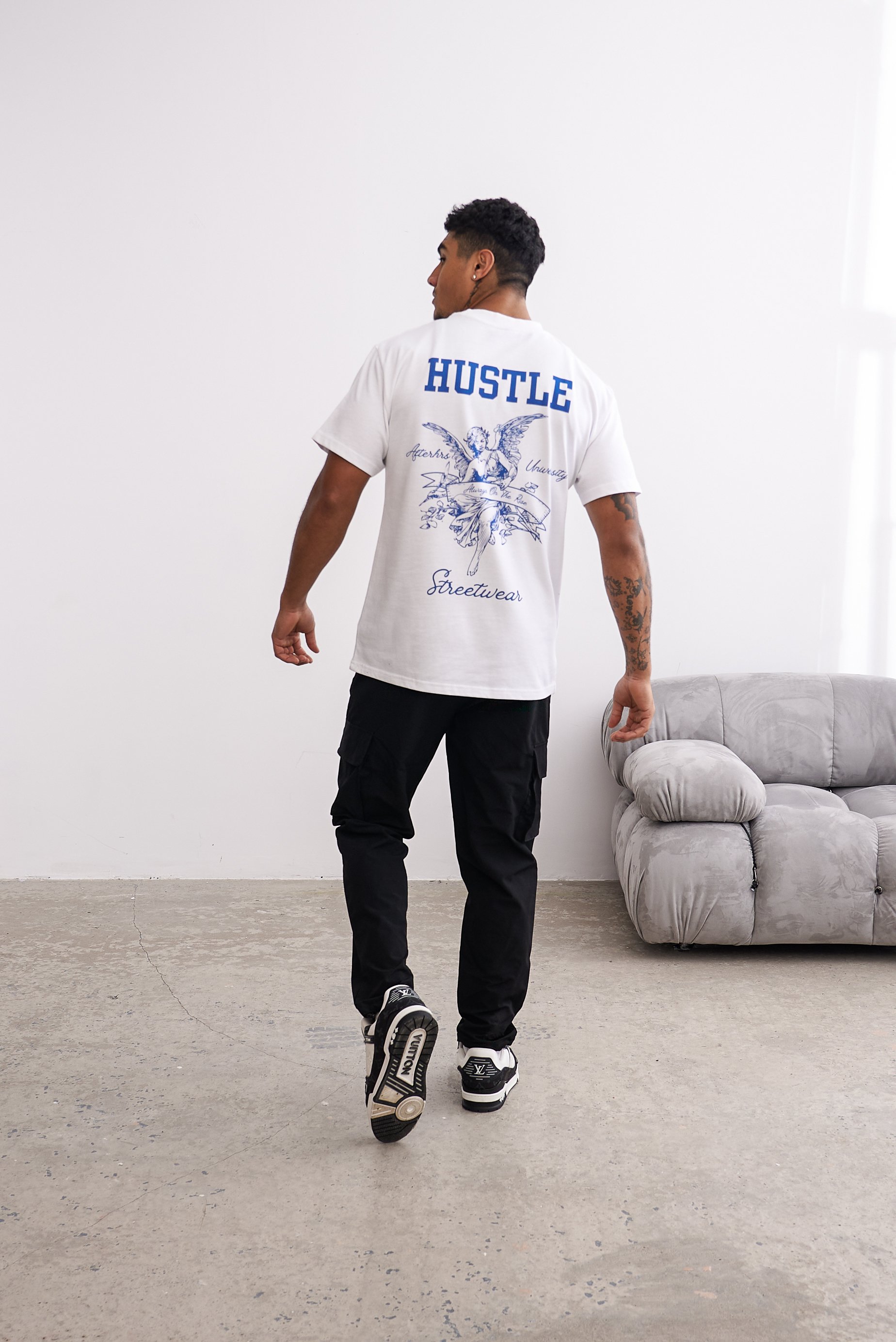 Man wearing a white T-shirt with "HUSTLE" text and graphics on the back, black cargo pants, and sneakers, walking away from a gray sofa.