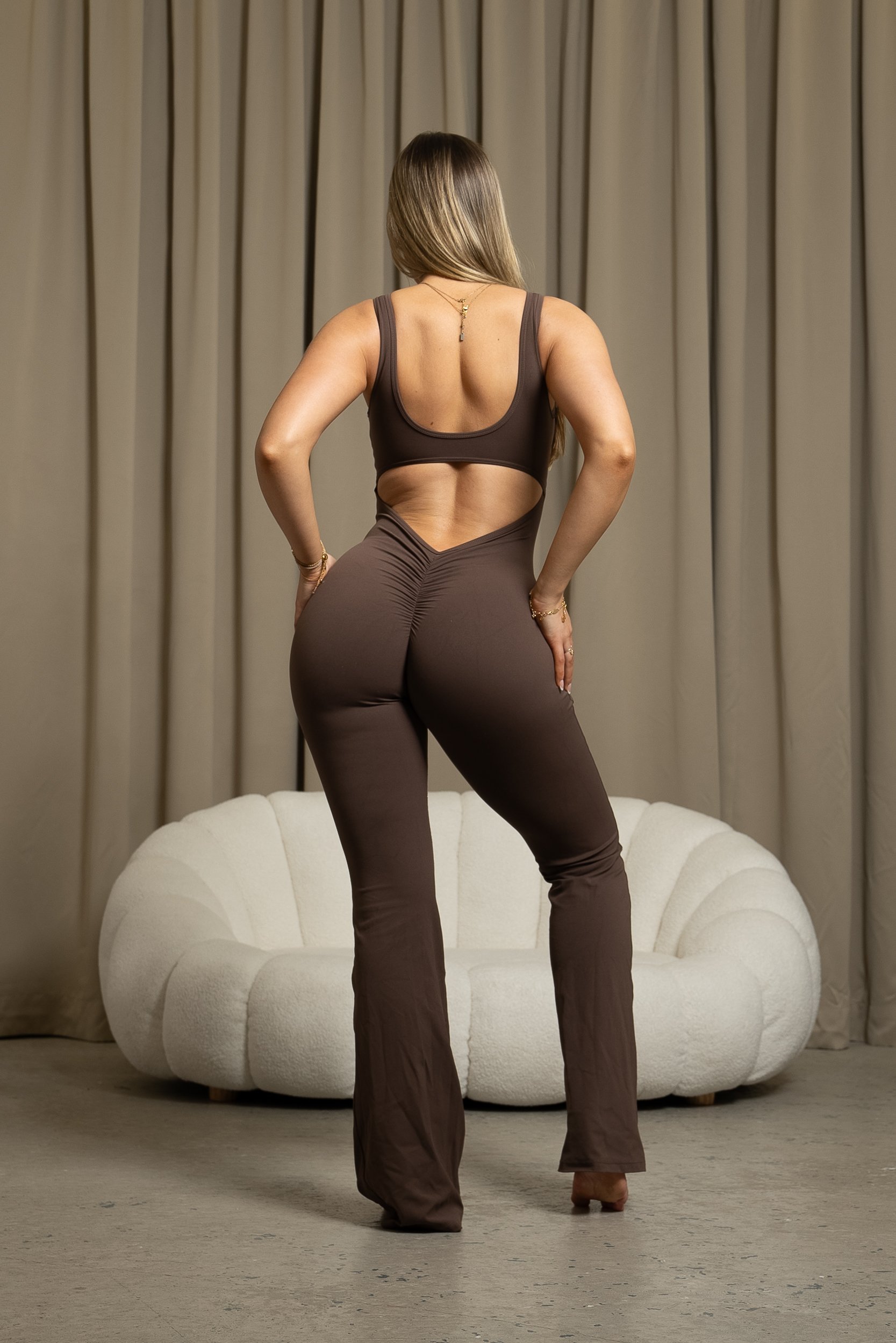Woman wearing a brown cutout jumpsuit standing in front of a white sofa and beige curtains.