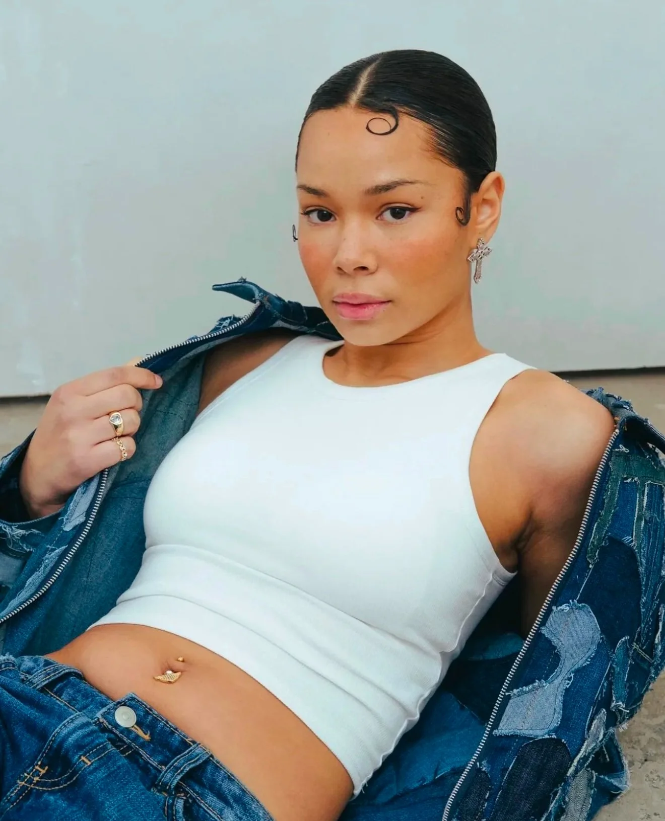 Person wearing a white tank top and denim jacket, with a belly button piercing and gold rings, posing confidently.