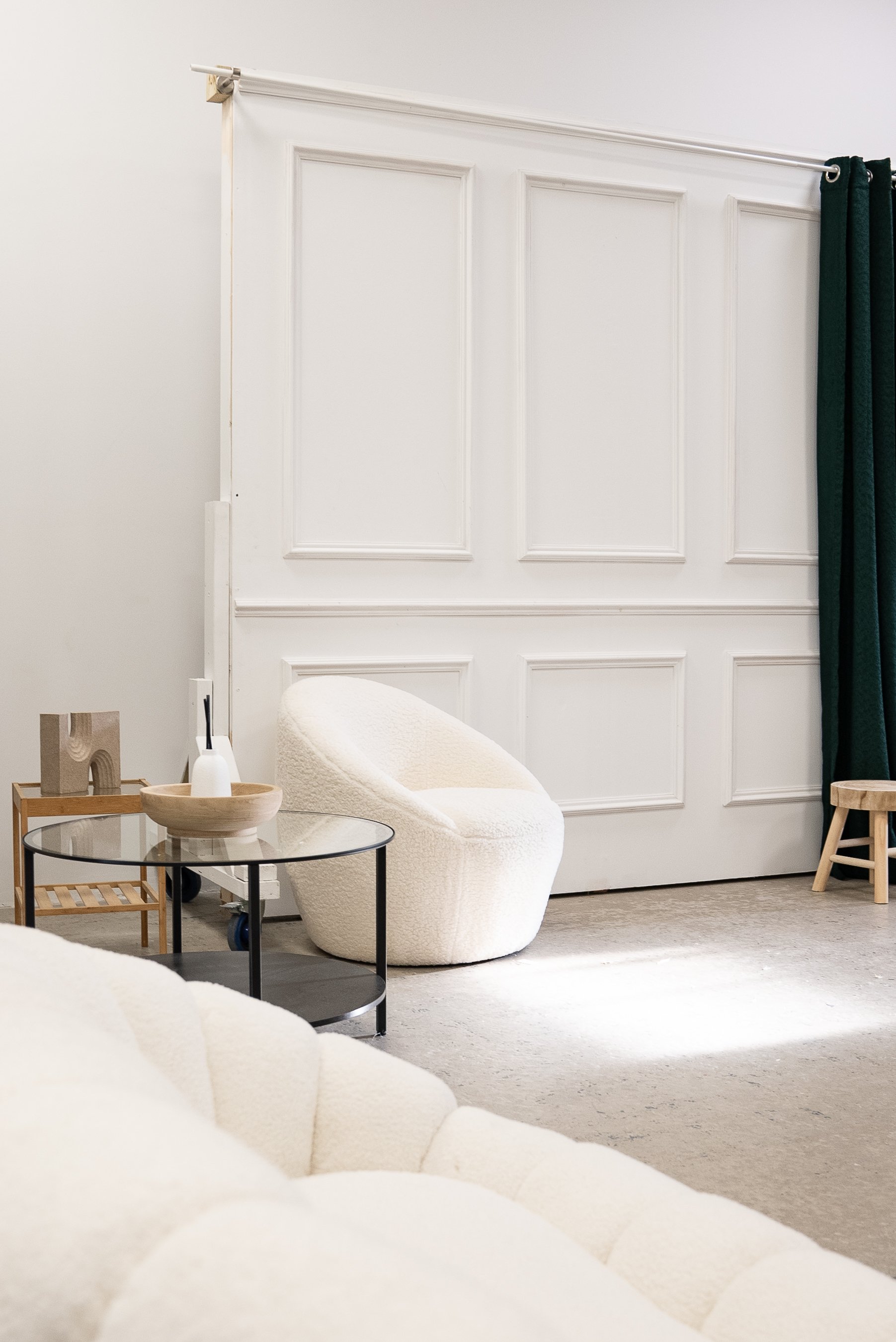 Modern living room with a white upholstered armchair, a round glass coffee table with a black metal frame, a wooden side table with decorative items, and panel walls. A dark green curtain and a beige sofa are partially visible.