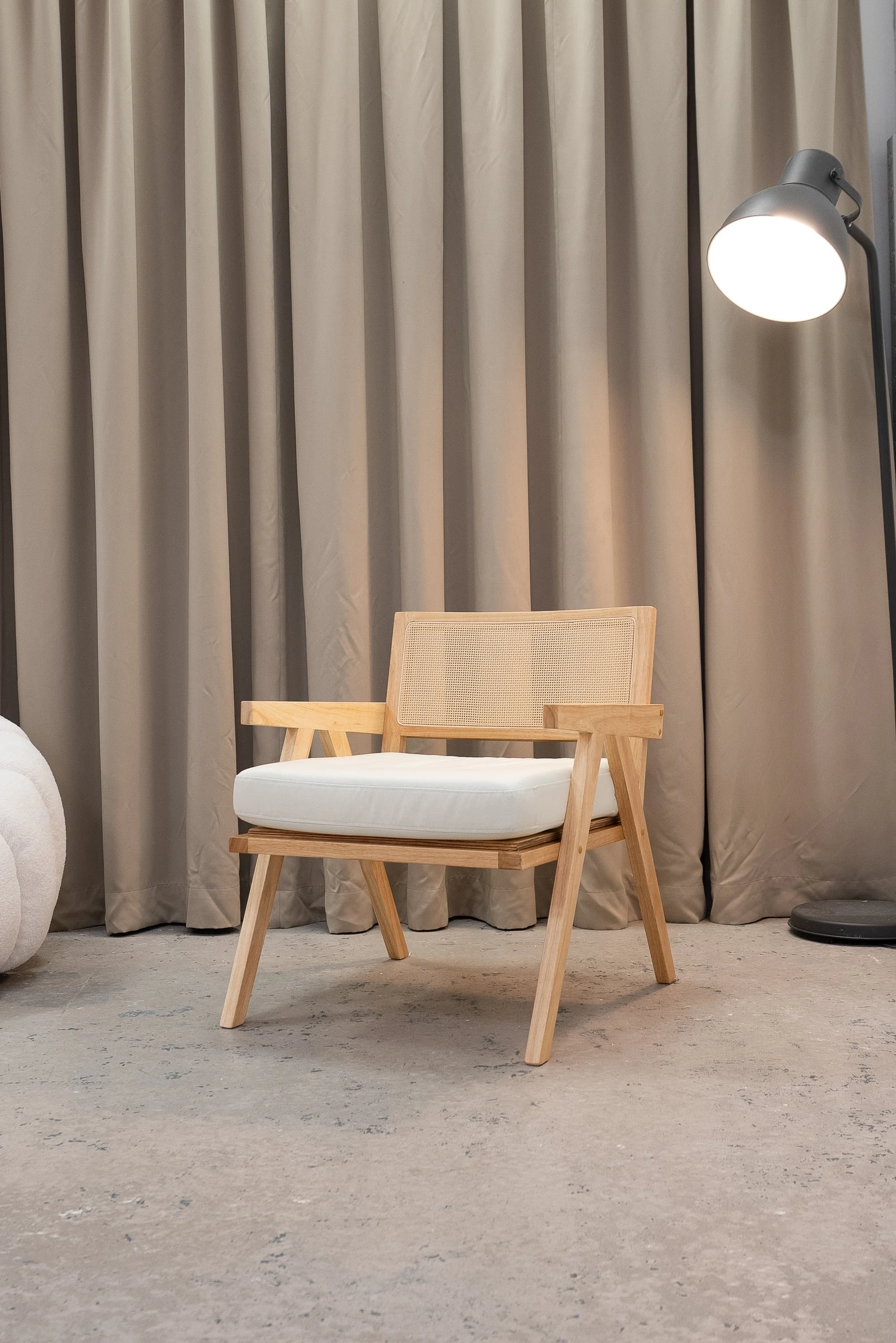 Modern wooden chair with white cushion, beside a floor lamp and beige curtains, on a concrete floor.