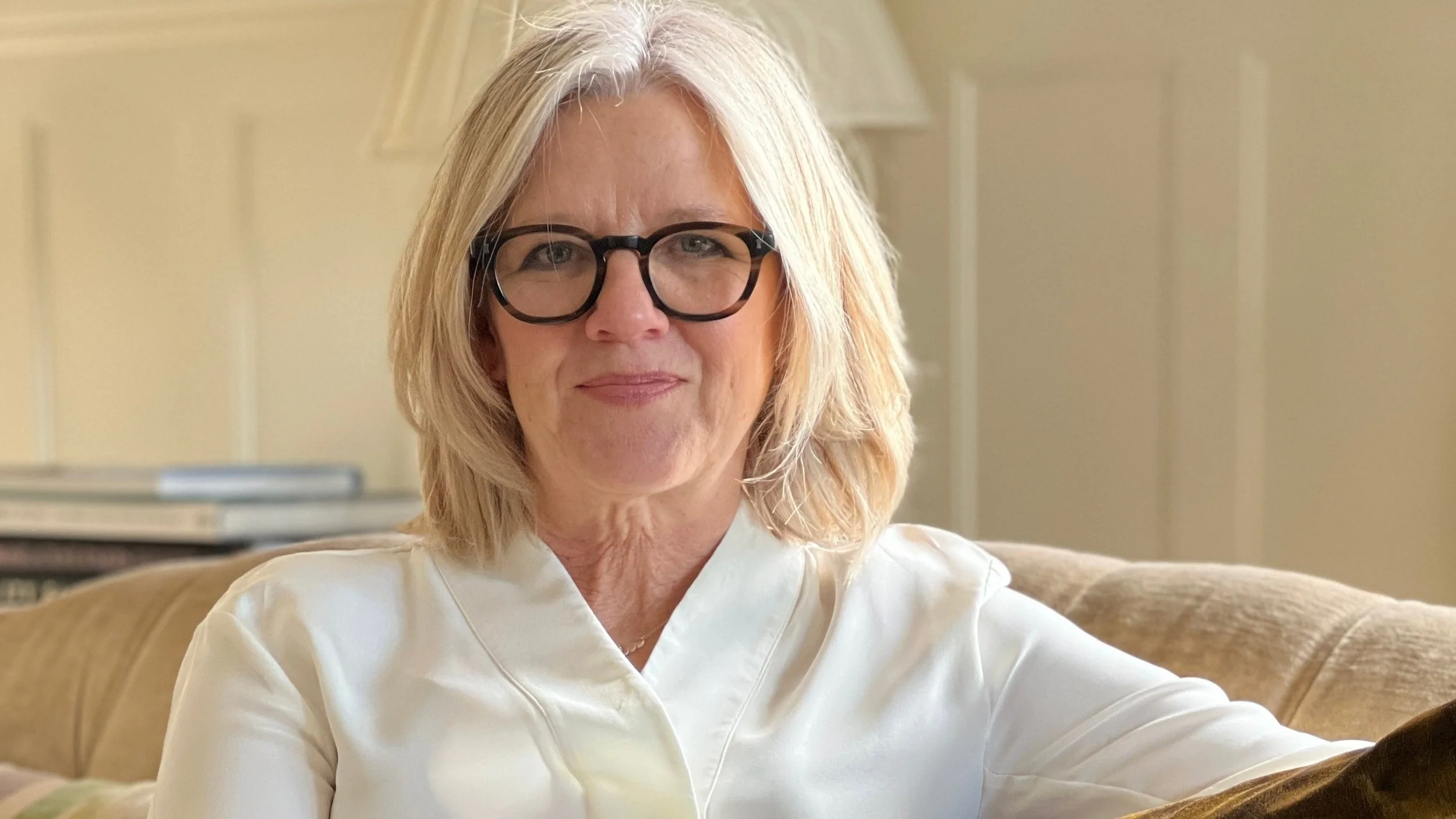 A middle-aged woman with blonde hair and glasses smiling while sitting on a couch in a cozy room.