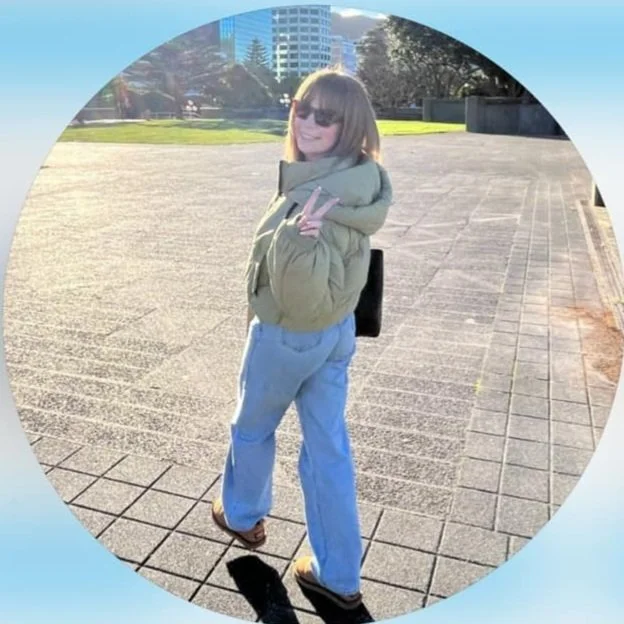 Person wearing sunglasses and a puffer jacket, giving a peace sign while walking on a paved area with greenery and buildings in the background.