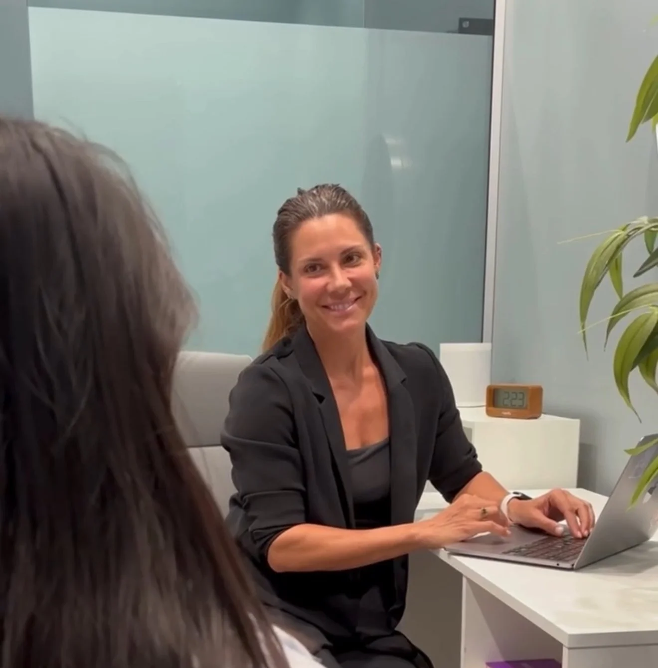 A woman with brown hair tied back, sitting at a desk with a laptop, smiling and talking to a person in the foreground.