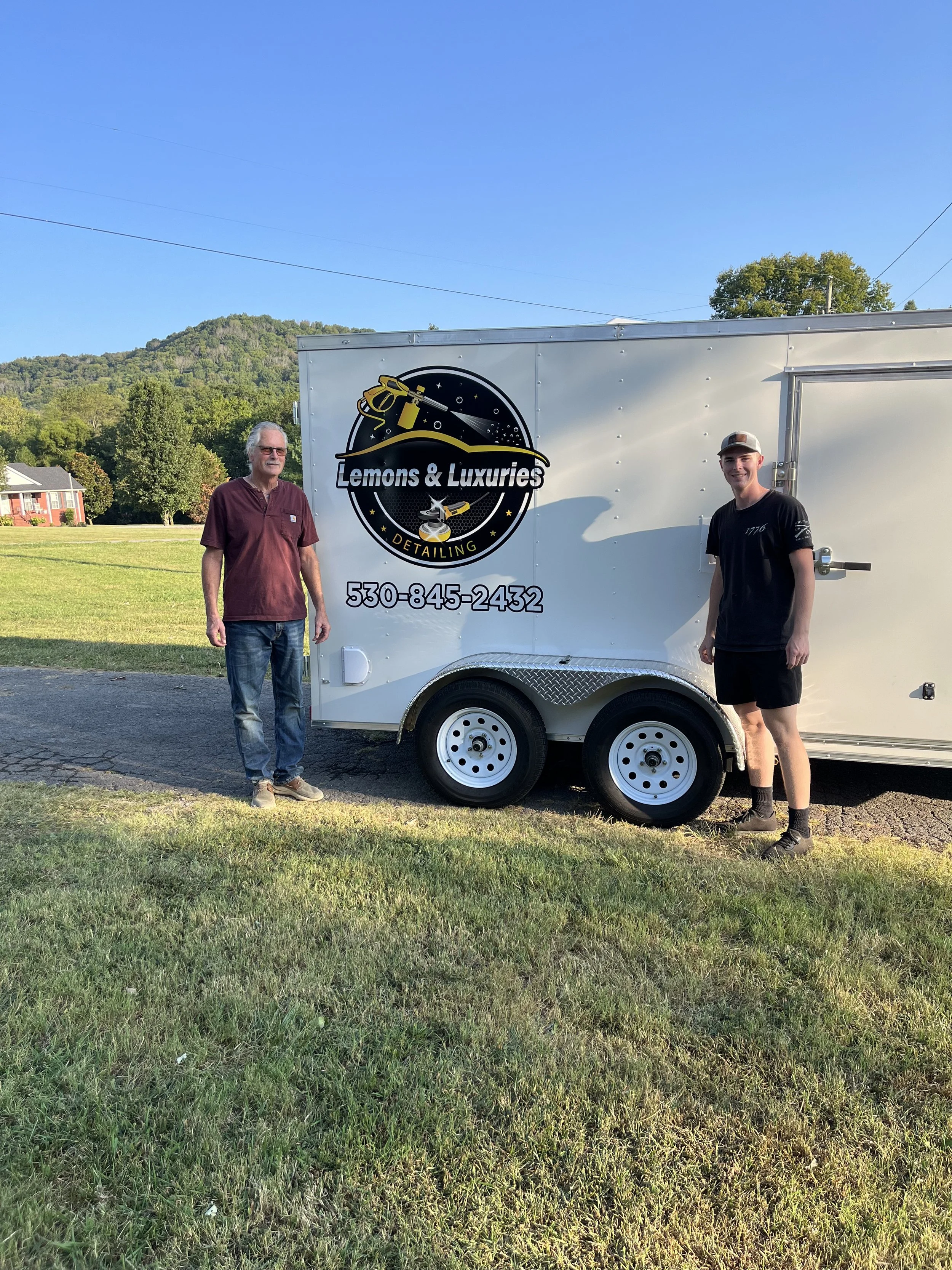 Two men standing in front of a white mobile detailing trailer with the 'Lemons and Luxuries Detailing' logo on it.