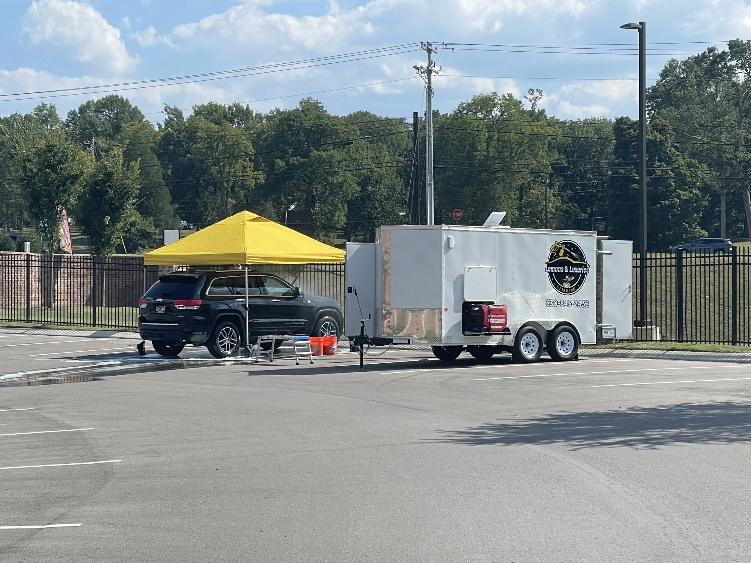 A mobile detailing trailer detailing a black Jeep SUV under a yellow canopy. The mobile detailing trailer is spraying the car from its 100 gallon water tank and is powered by a 5000 watt generator.