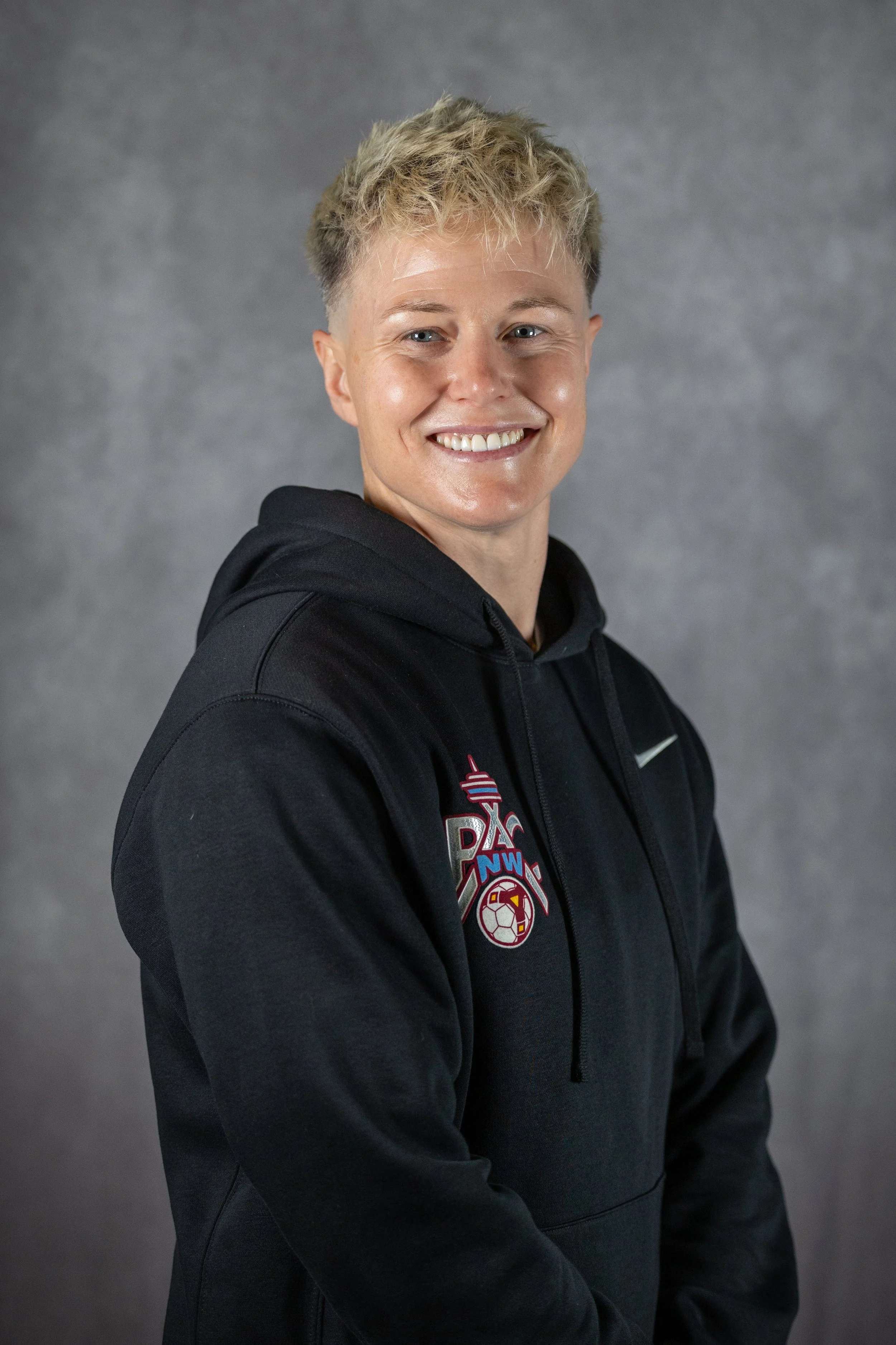 Young woman smiling, wearing a black hoodie with a sports logo on the chest, against a plain gray background.