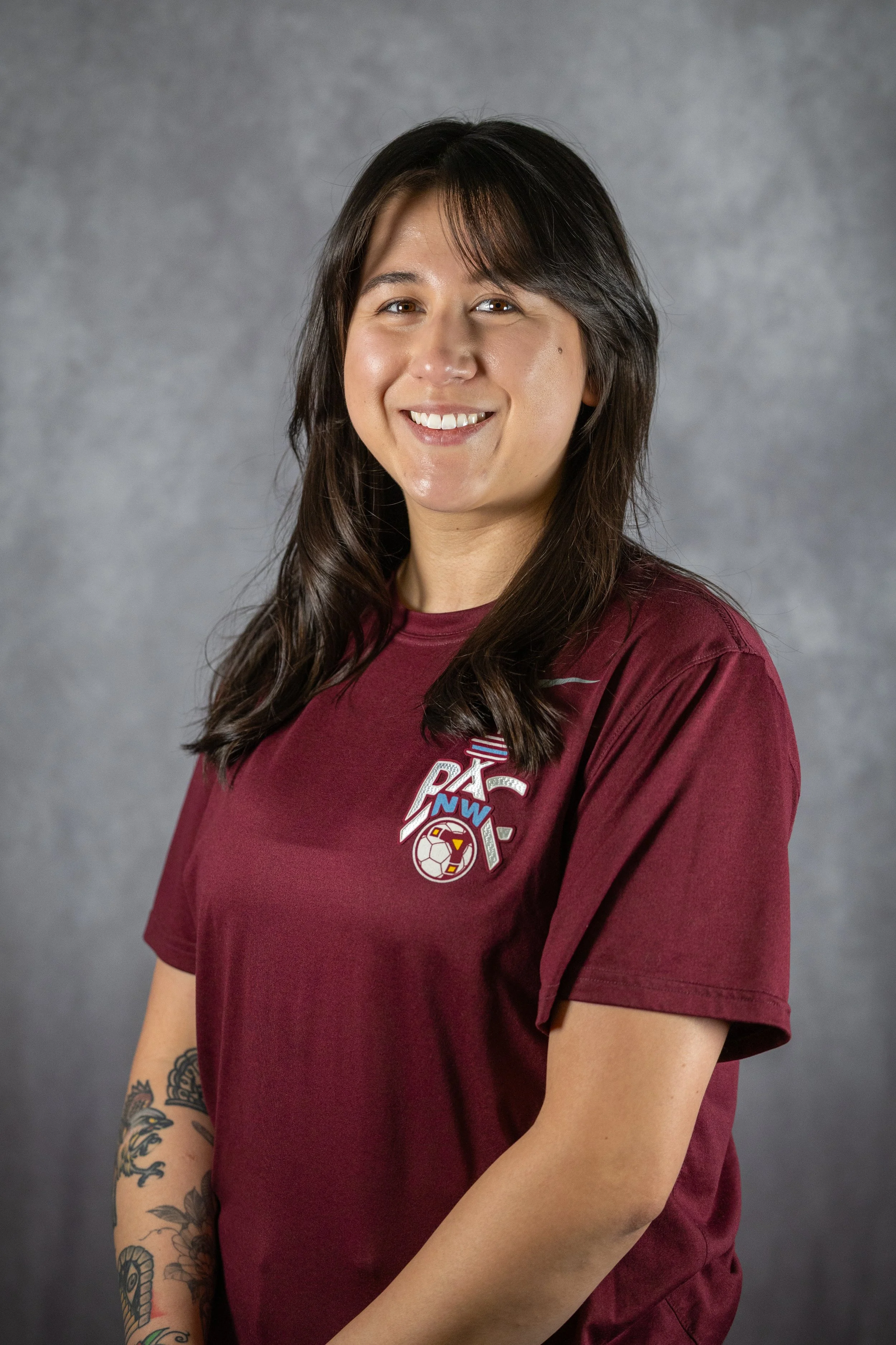 A young woman with dark brown hair and tattoos on her left arm, wearing a maroon t-shirt with a sports logo, smiling against a gray background.