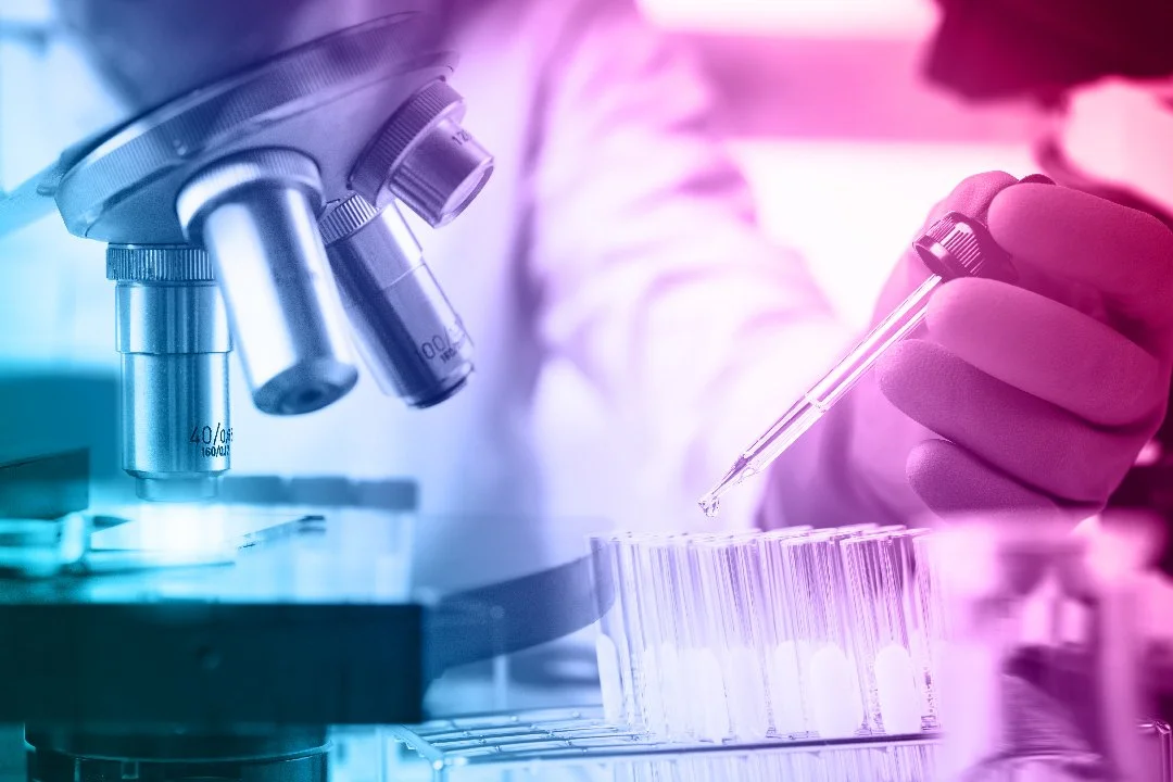 Closeup of microscope lenses, alongside a rack of test tubes, and a scientist's gloved hand holding a dropper.