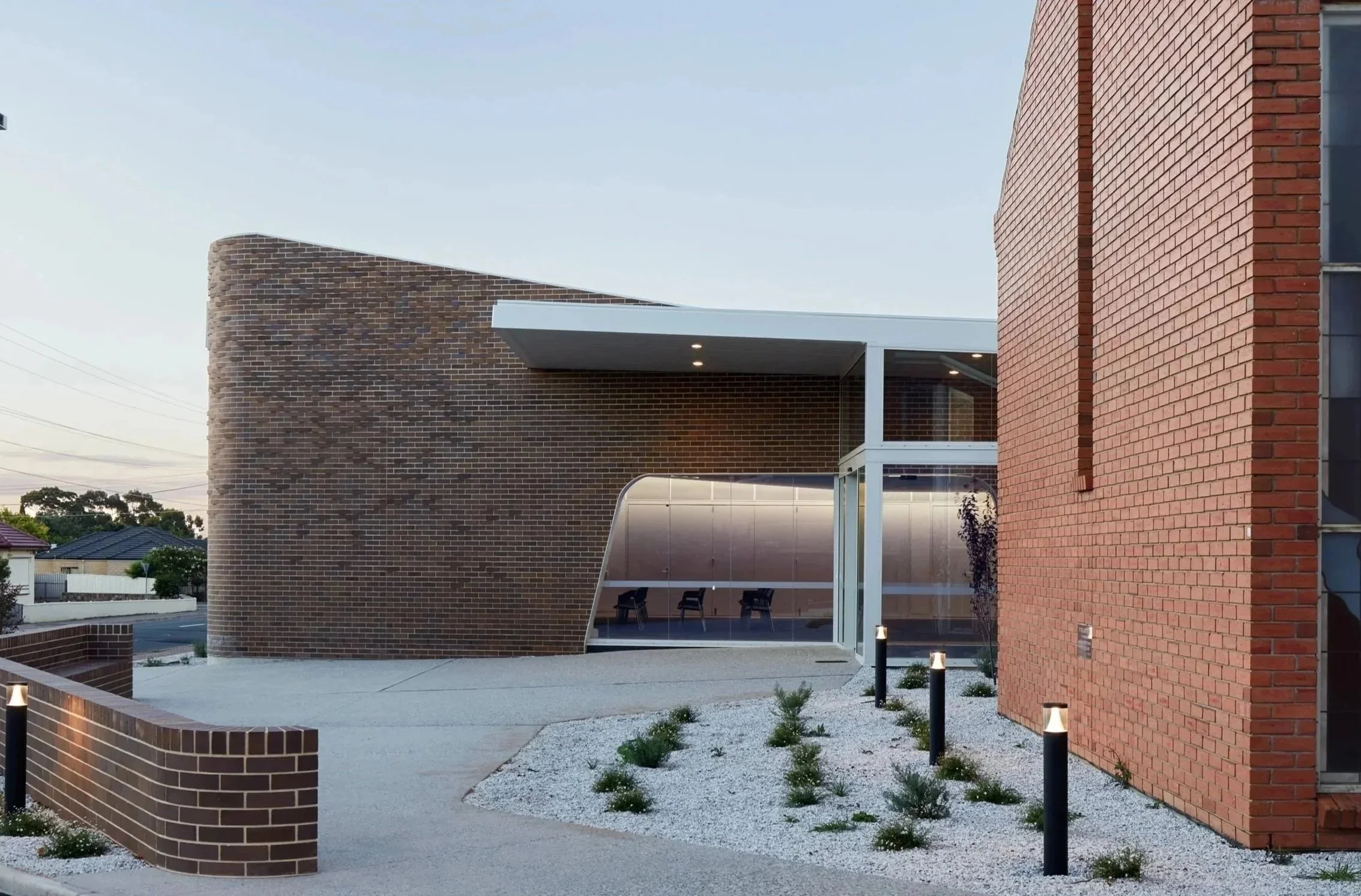 Modern building with brick walls, large windows, and an outdoor seating area with chairs, surrounded by small plants and pathway lights.