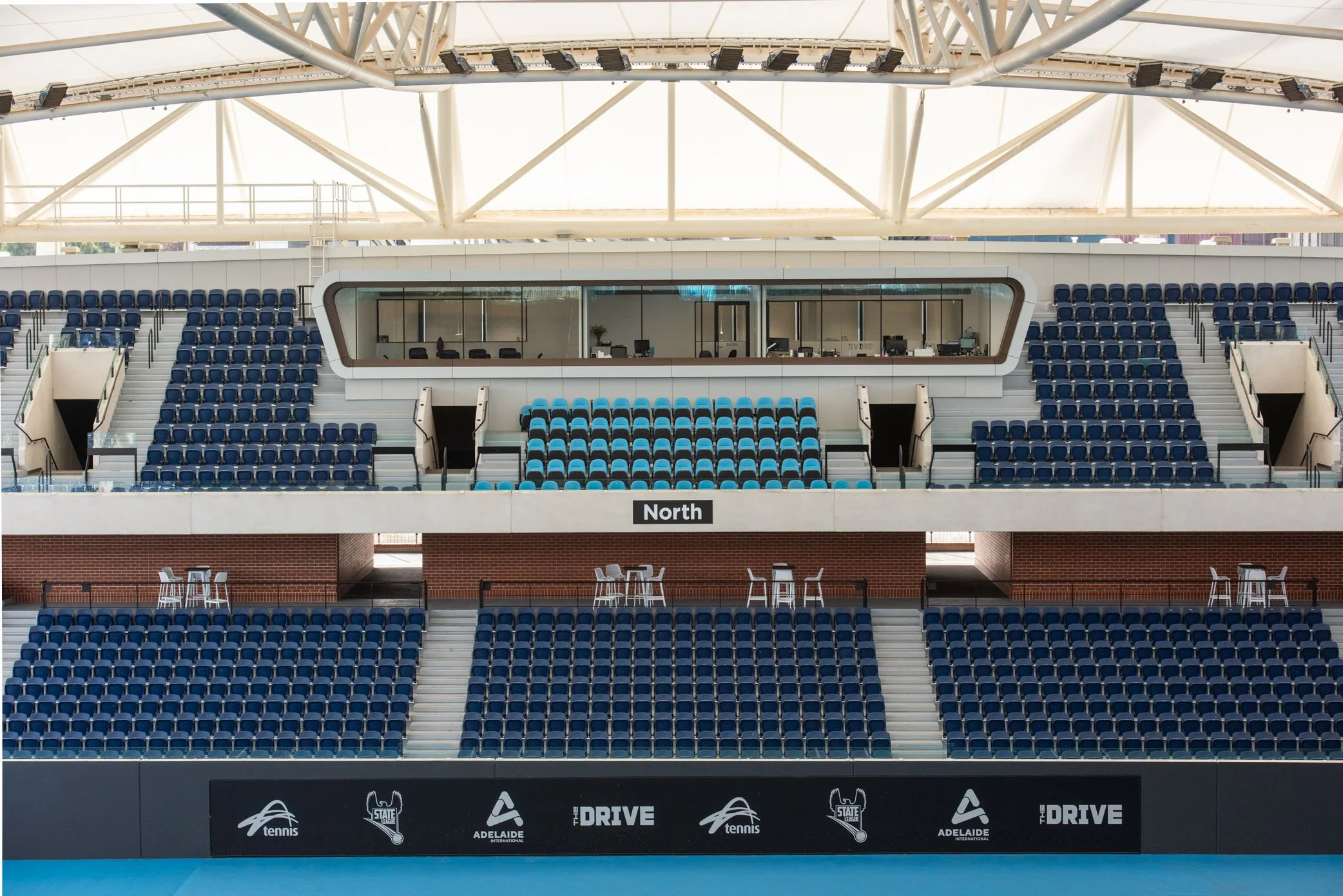 Empty stadium seating area with blue chairs, a glass-enclosed press box, and a black banner featuring event logos and text.