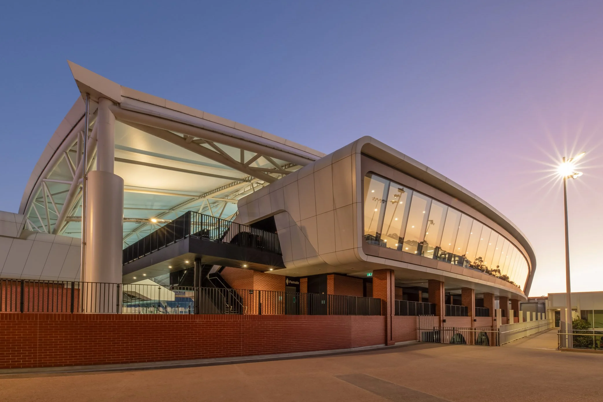 Modern building with a curved glass facade and metal support structures, illuminated inside, with a brick base and a fence, under a clear evening sky with the sun setting and a streetlight turned on.