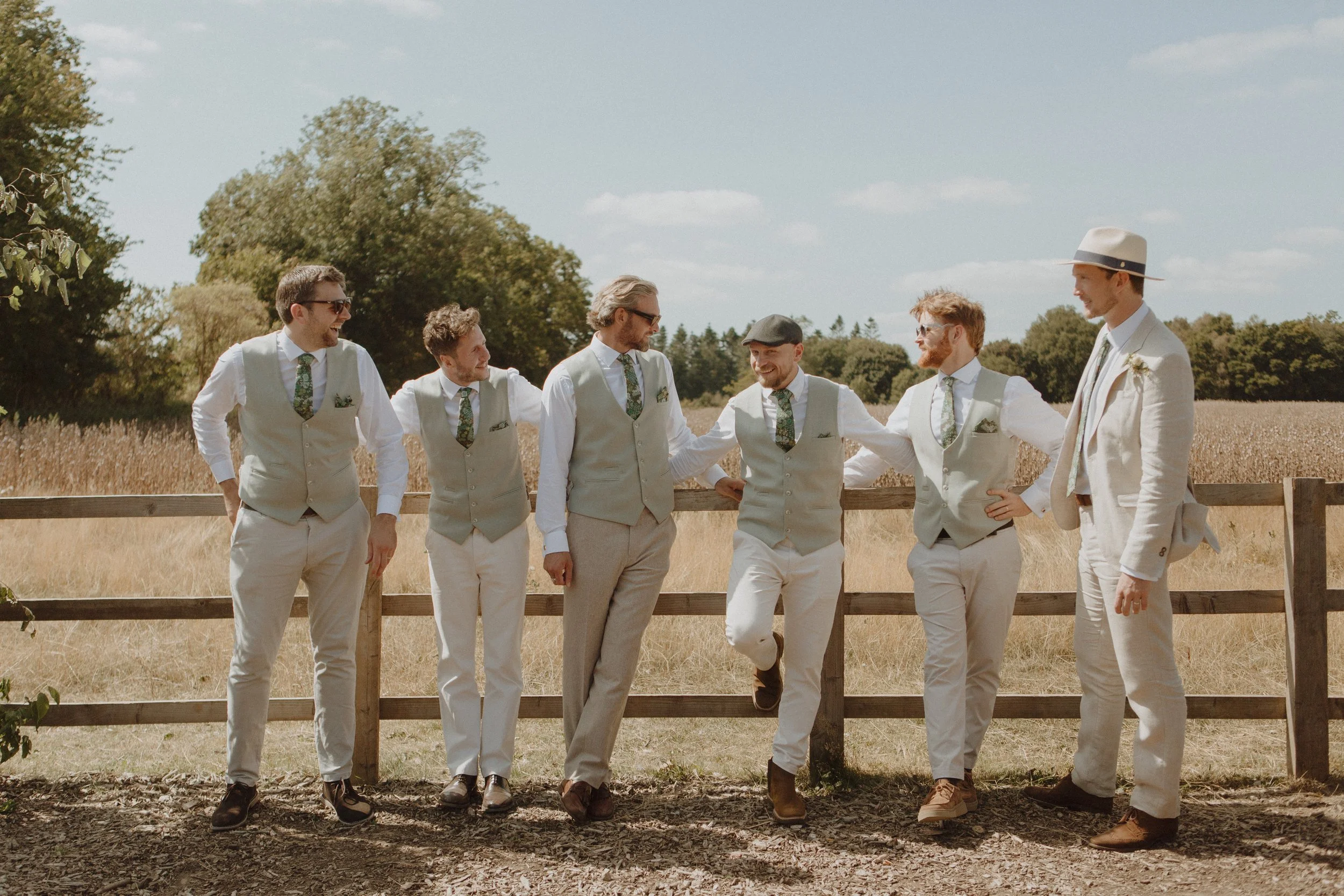 A group of seven men dressed in vintage-style clothing, standing outdoors by a wooden fence, smiling and chatting on a sunny day in a rural setting.