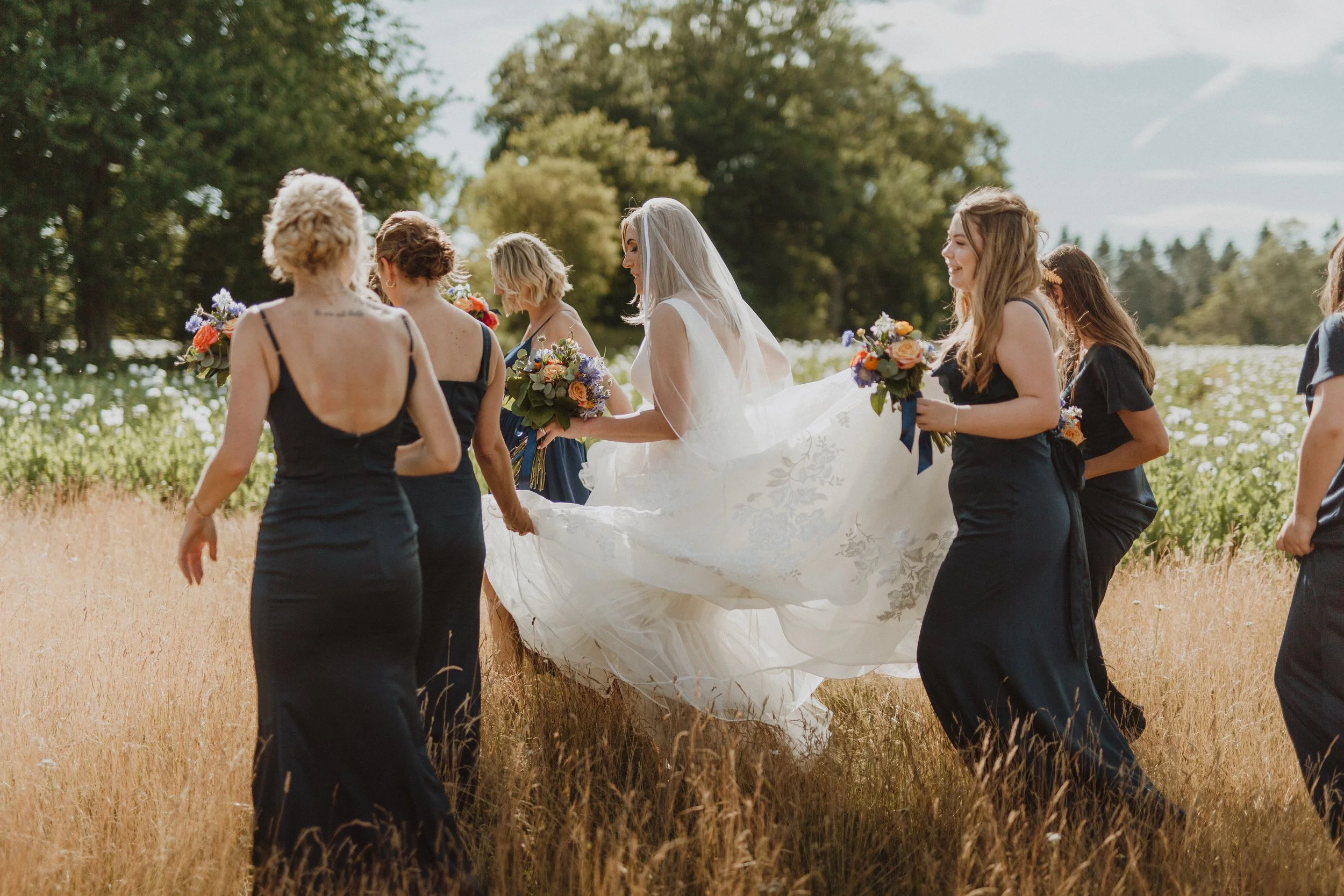 A bride in a white wedding dress and veil walking through a field, surrounded by bridesmaids in navy dresses holding bouquets of flowers.