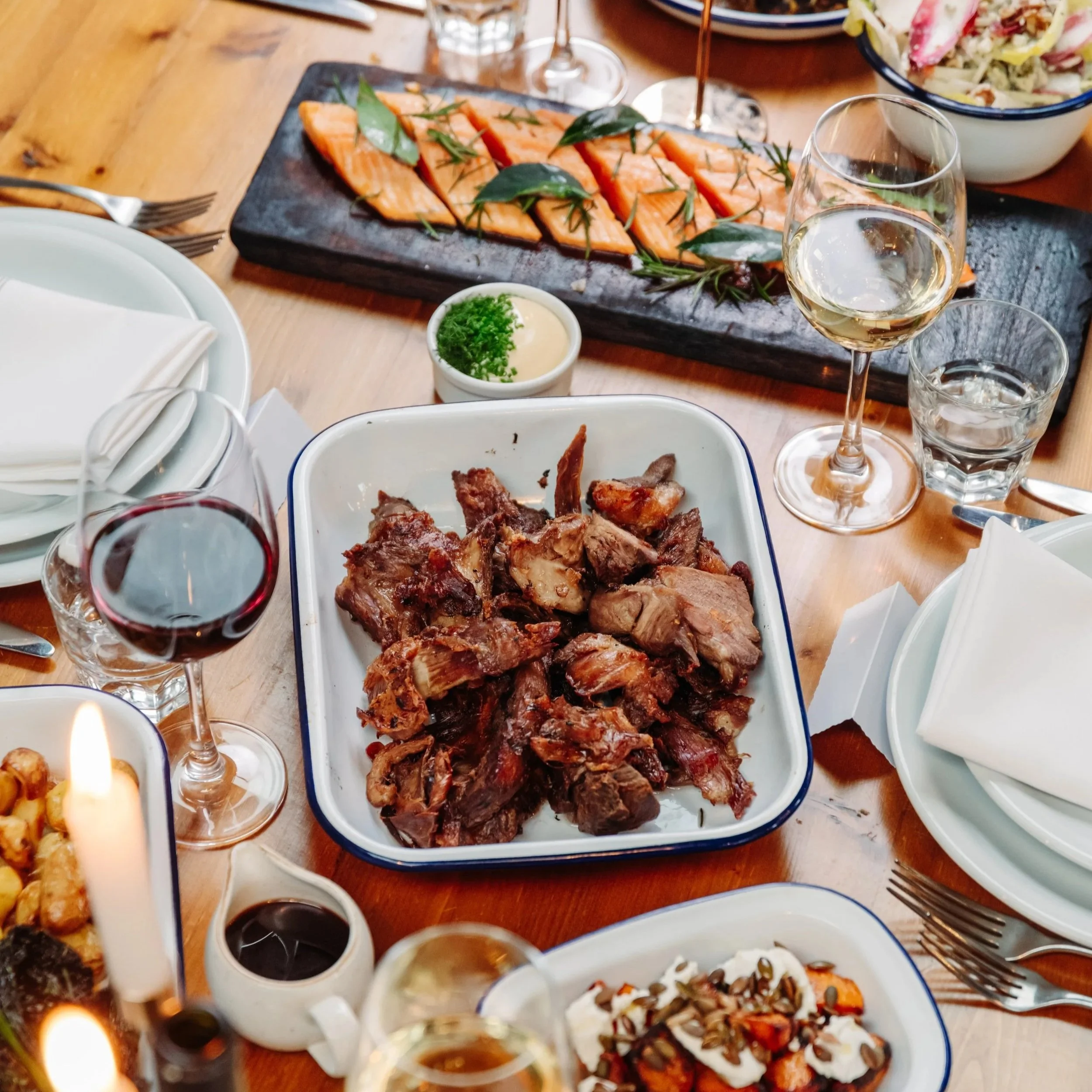 A table with various dishes and drinks, including a platter of roasted meat, a bowl of salad, a slate board with sliced salmon garnished with herbs, a glass of white wine, a glass of red wine, and several empty plates and utensils.