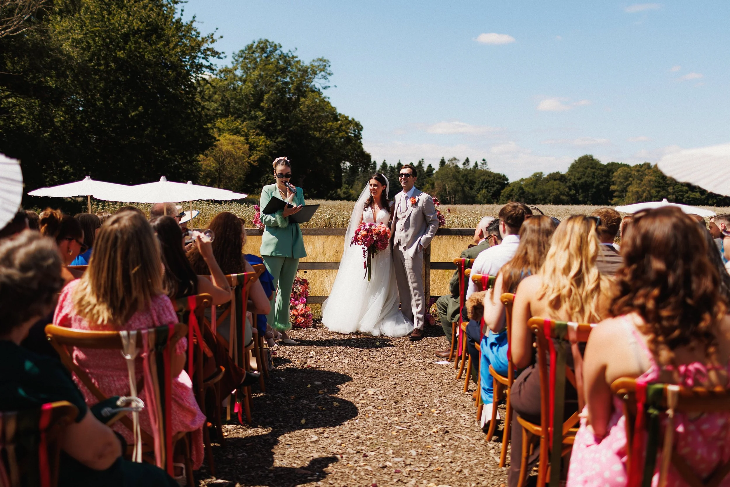 A wedding ceremony outdoors with a bride and groom standing in front of a seated guest audience during daytime. The bride is in a white wedding gown holding a bouquet of pink flowers, and the groom is in a light gray suit. An officiant, standing to the left, appears to be reading from a book. There are white umbrellas and lush trees in the background.