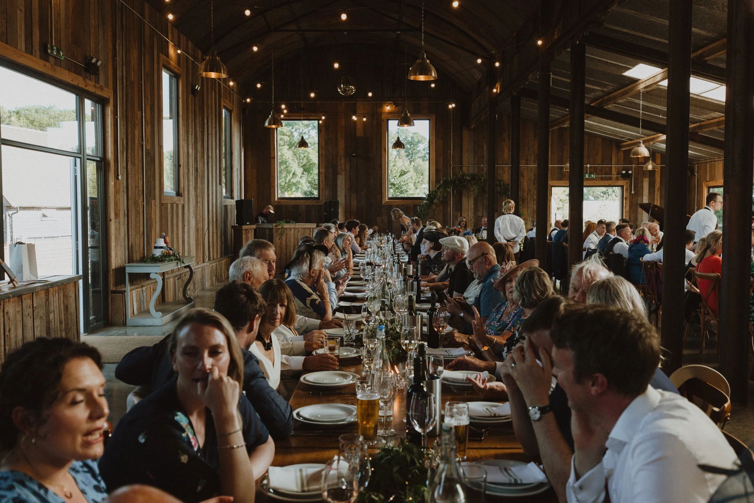 People dining at a long table in a rustic wooden banquet hall with string lights overhead.