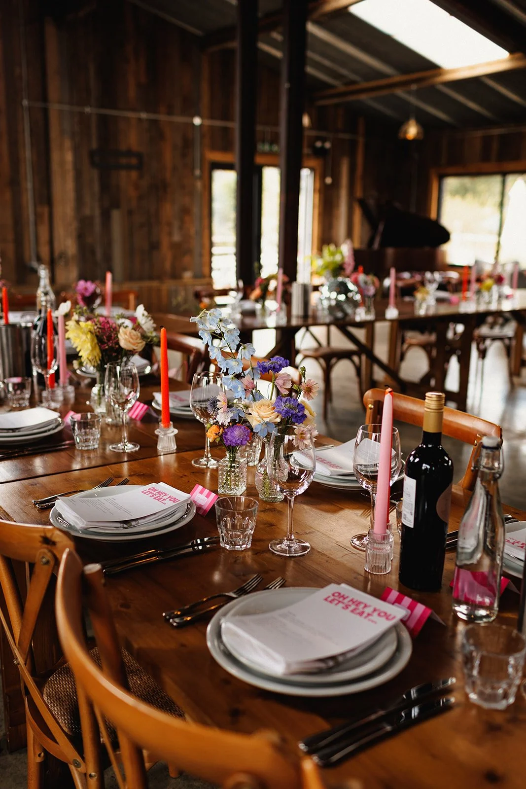 A rustic wooden dining table set for a celebration with floral centerpieces, pink candles, wine bottles, and place settings that include plates, glasses, utensils, and pink-striped napkins. From Jason Williams Photography