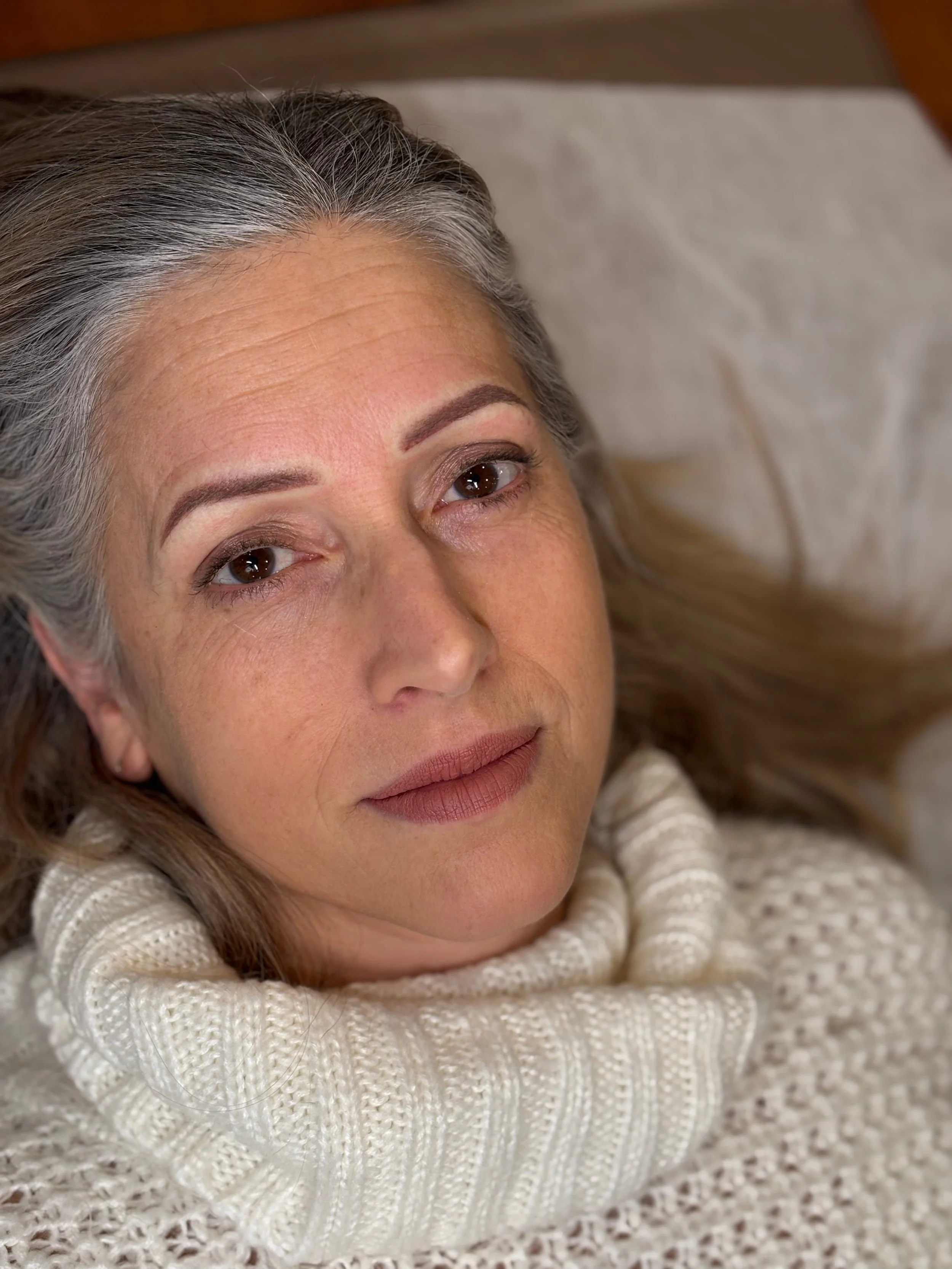 image of 50 year old beautiful woman with long gray hair, large brown eyes and beautiful berry matte lip color and fresh powder brows , women wearing a cream turtleneck sweater laying back looking into camera , up close image of woman