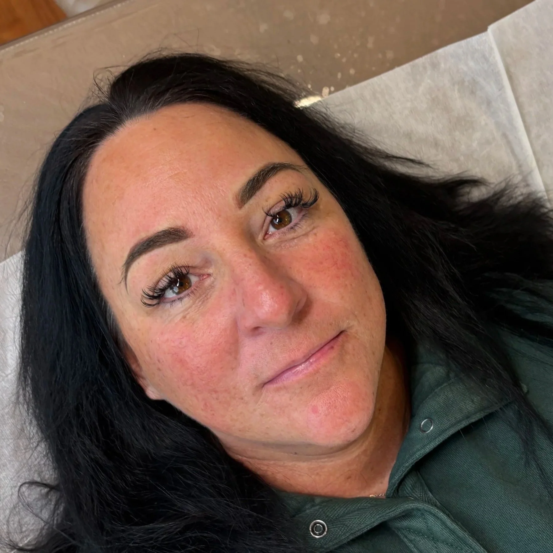 Italian womens face up close with a green shirt on, black long straight hair, long false eyelashes, brown eyes and dark brown brows with powder brow permanent makeup