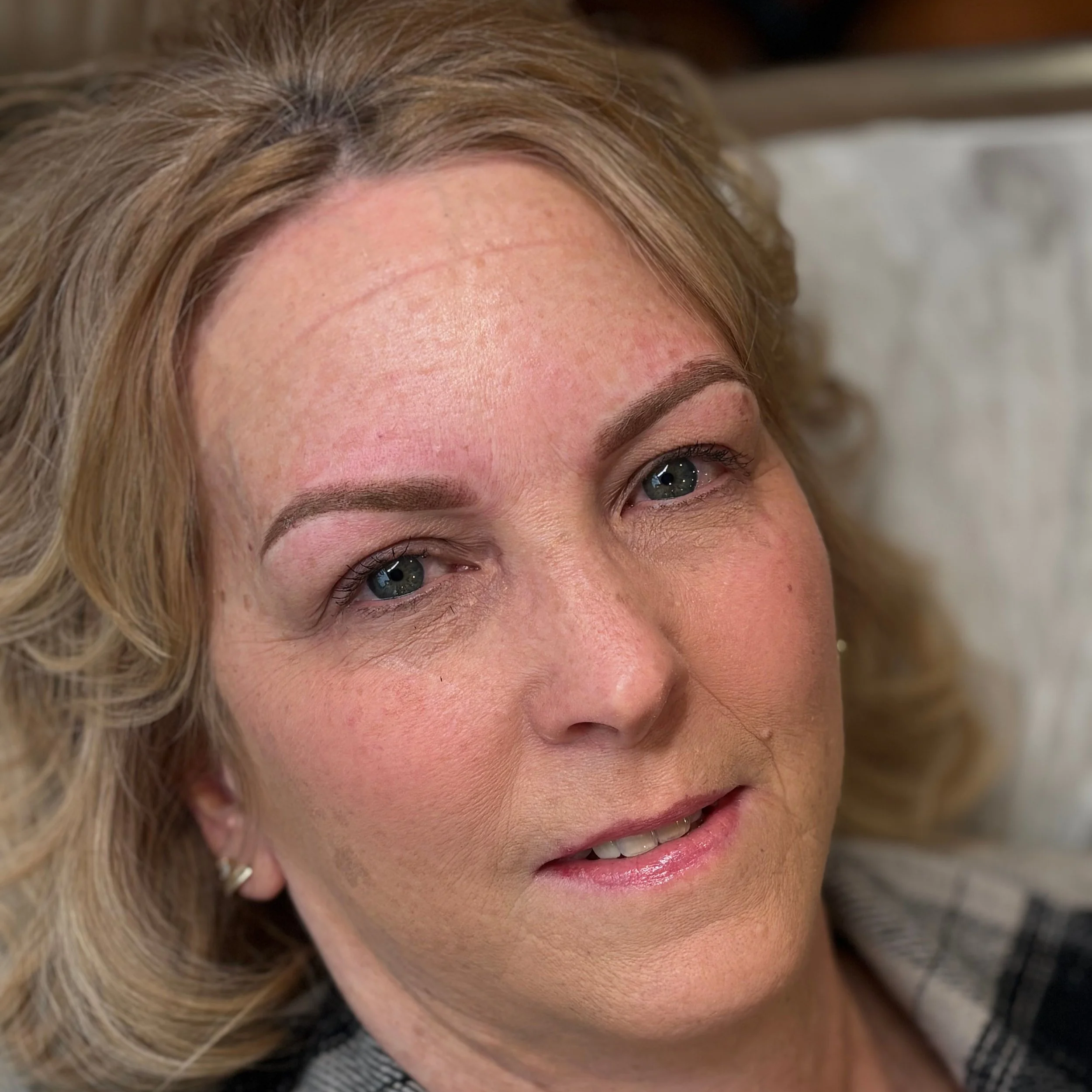 older lady with blonde soft curls , with small lips and bright blue eyes wearing a flannel shirt, lady laying back looking at camera, lady with permanent makeup , blonde woman with ash brown eyebrows