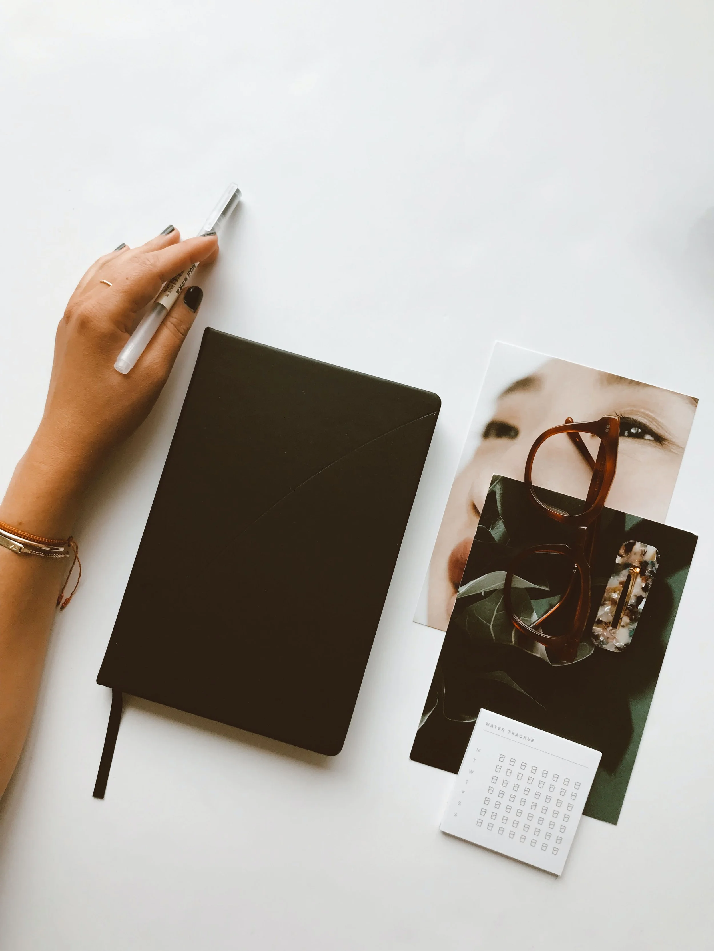 A white desk with a person's hand holding a white pen, a closed black notebook, two photographs of a person's face and a person's eye with glasses, a small water tracker calendar, and a pair of tortoise shell glasses.