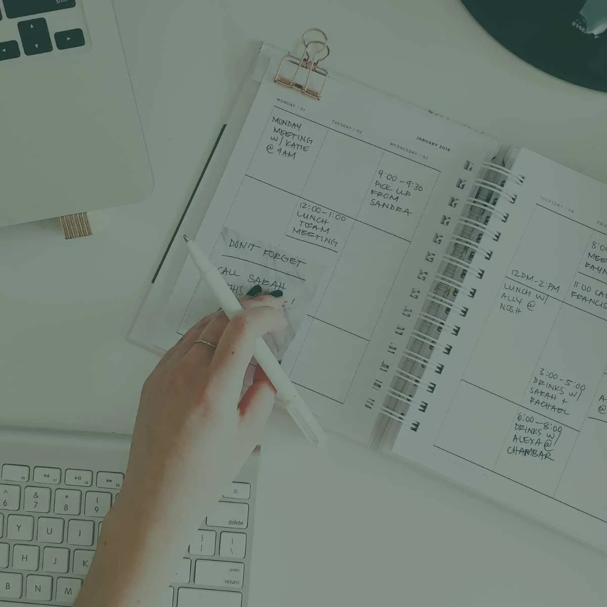 A weekly planner with handwritten notes on a white desk, partially visible laptop keyboard, and a person in the process of writing with a white pen.