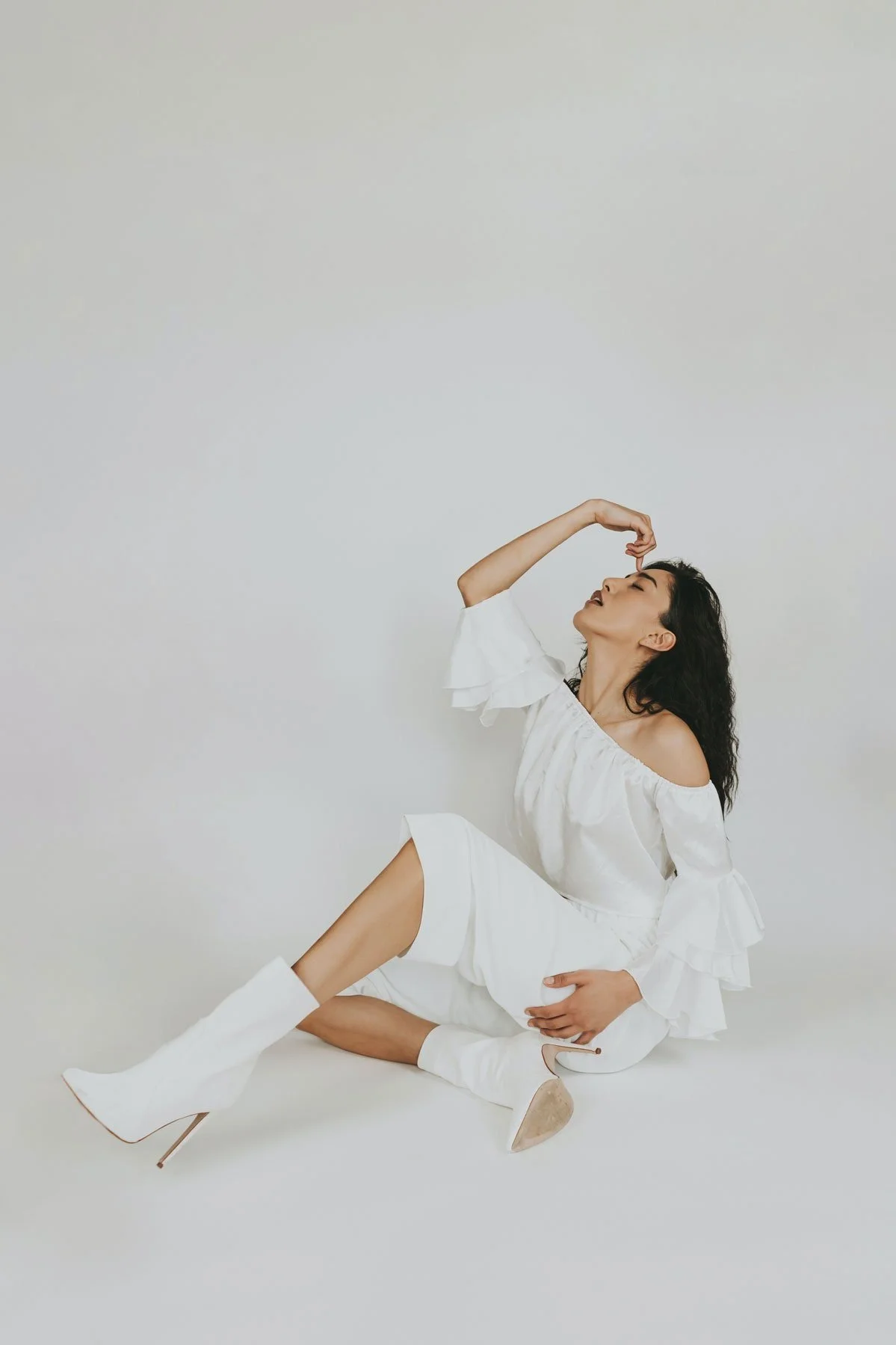 A woman with long dark hair, wearing an off-the-shoulder white dress and white high-heeled boots. She is sitting on the floor against a plain white background, with her eyes closed and her hand touching her forehead.
