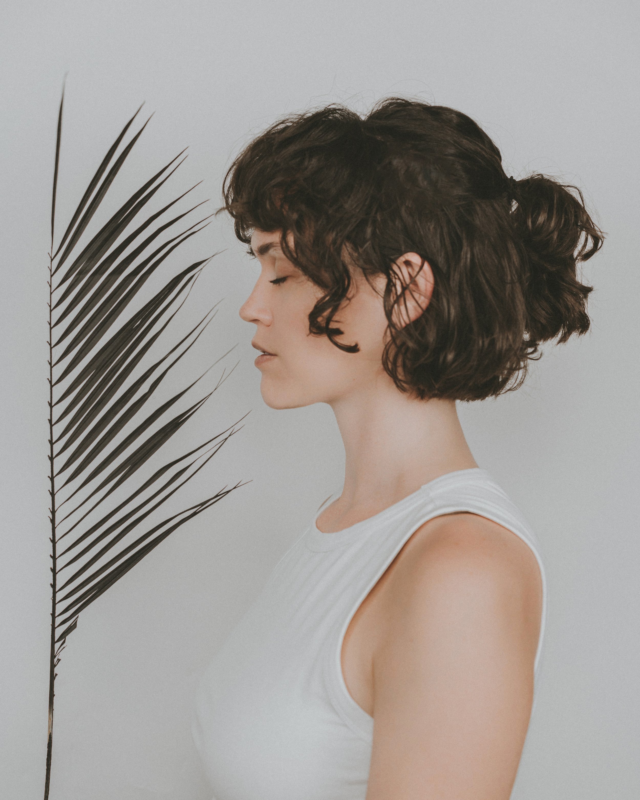 Profile of a young woman with short, curly brown hair, closed eyes, and light skin, standing next to a dried palm leaf against a plain light gray background.