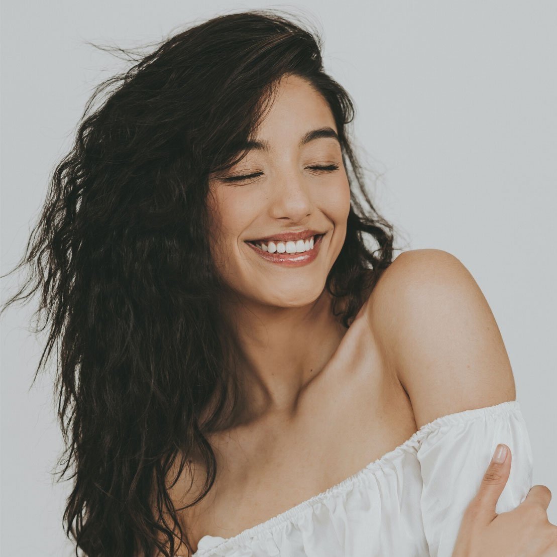 A woman with curly hair smiling with eyes closed, wearing a white off-shoulder top.