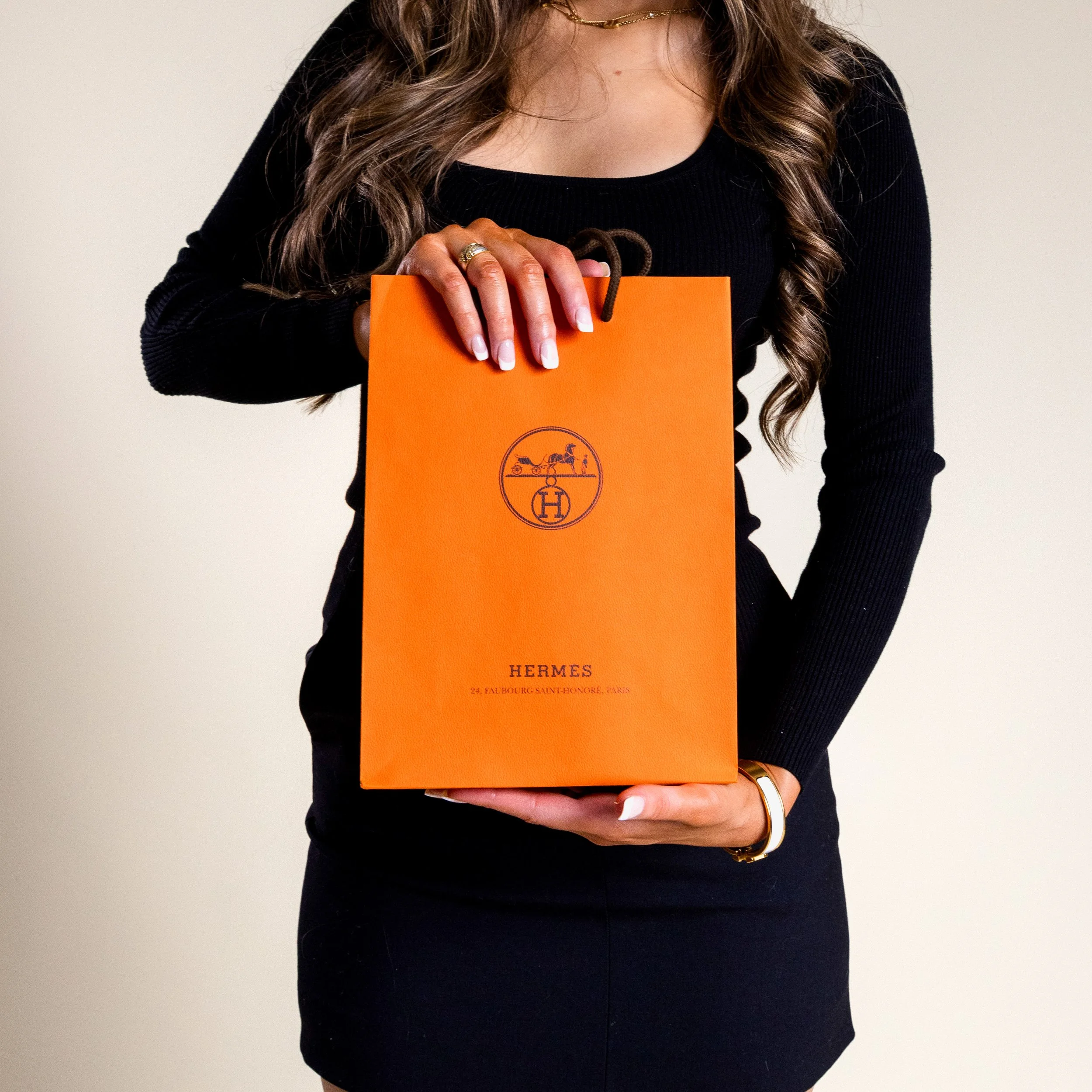 A woman holding an orange Hermès shopping bag with black handles, wearing a black long-sleeve top and dark skirt, with wavy hair and jewelry.