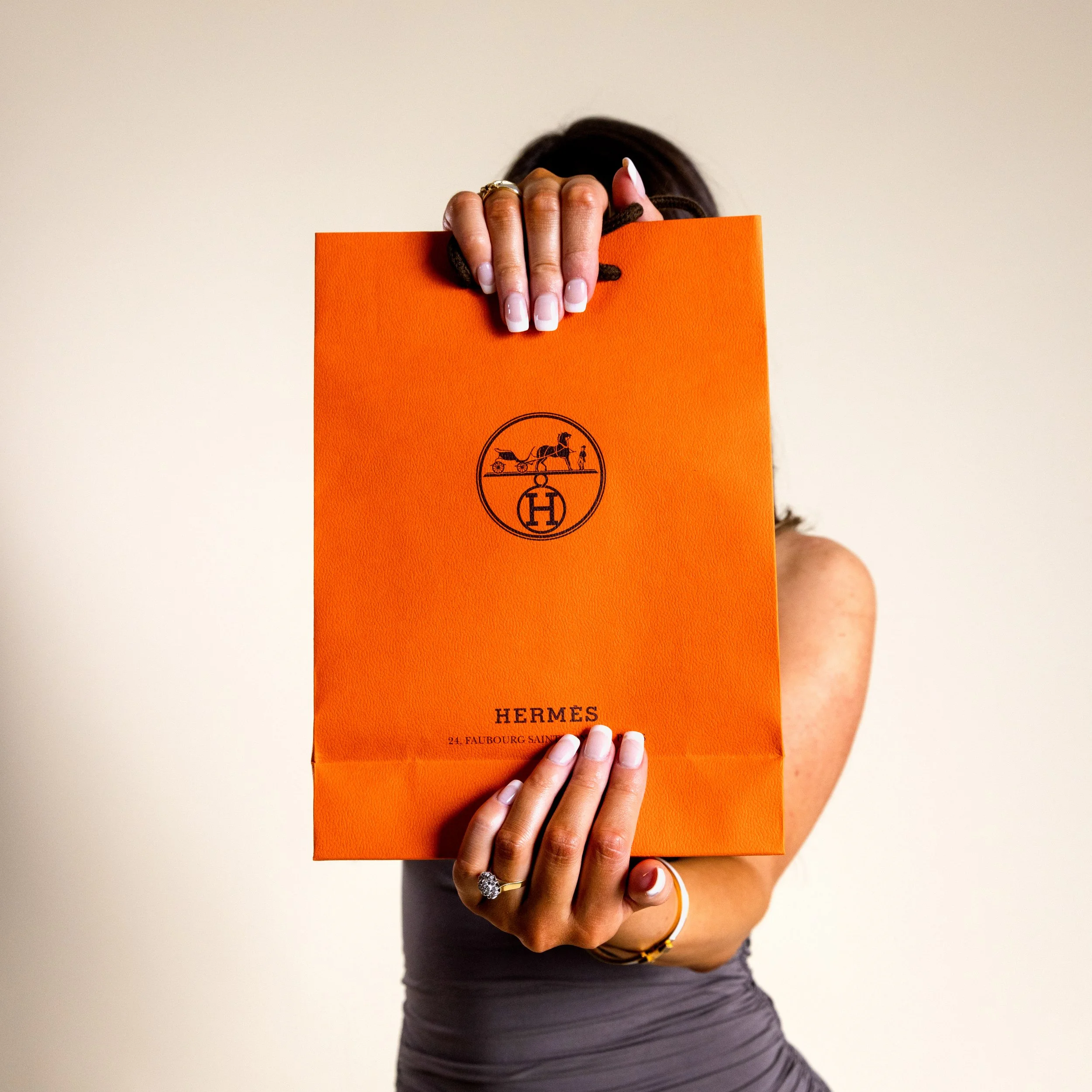 A person holding an orange Hermès shopping bag in front of their face, obscuring it from view, with a plain background.