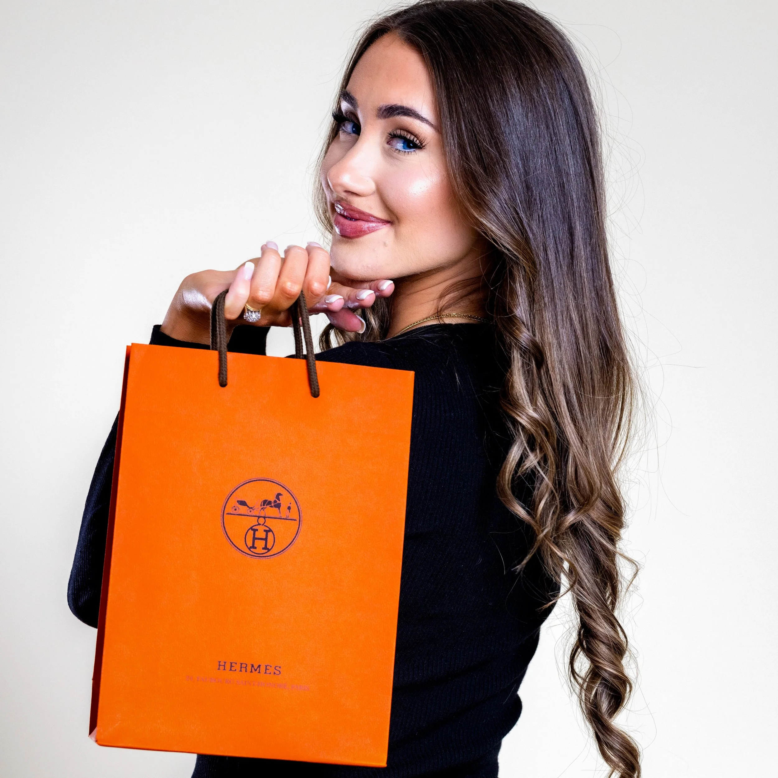 A woman with long wavy hair holding an orange Hermès shopping bag, smiling and looking over her shoulder.