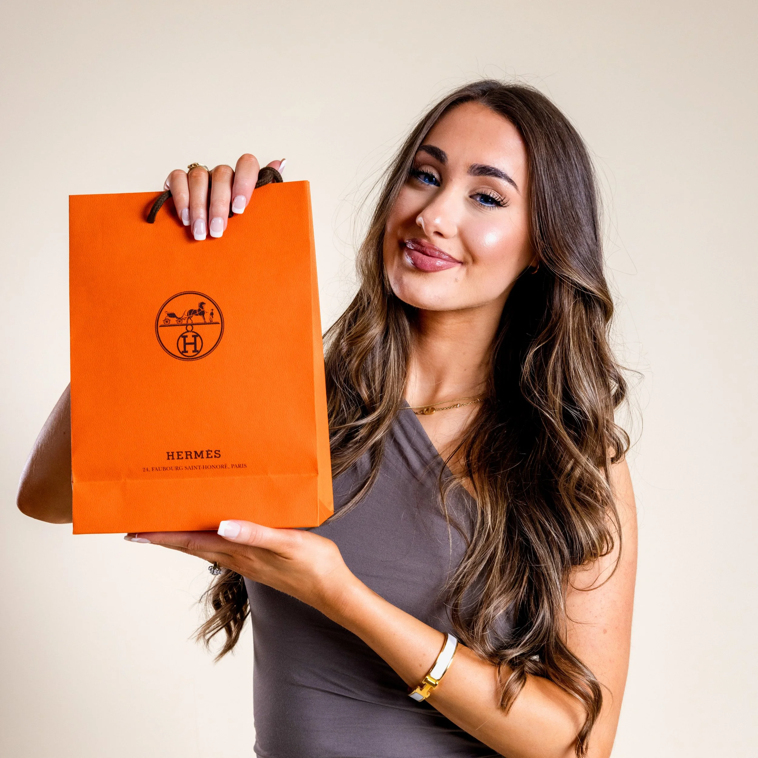 A woman with long brown hair holding an orange Hermès shopping bag smiling at the camera.