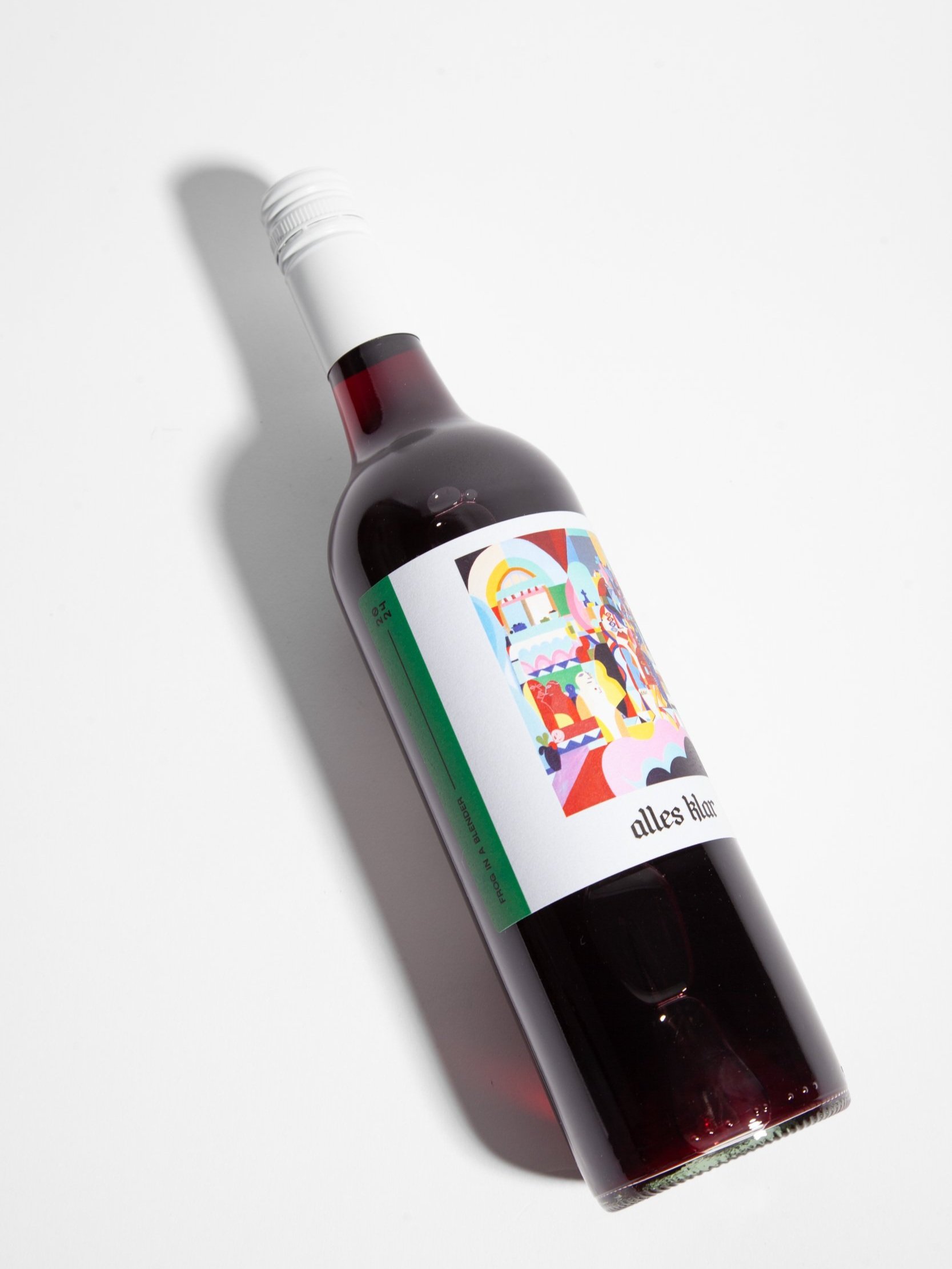 A single bottle of Alles Klar Frog In A Blender 2024, a full-flavoured chilled red, is shown lying diagonally on a white surface, casting a soft shadow to its left.