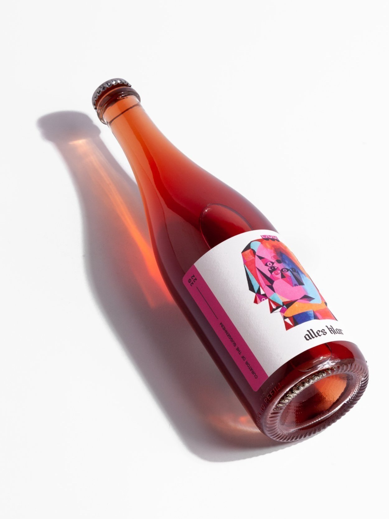 A single bottle of Alles Klar Curator of the Guggenheim Pet Nat, a sparkling rosé wine, is shown lying diagonally on a white surface, casting a soft shadow to its left.