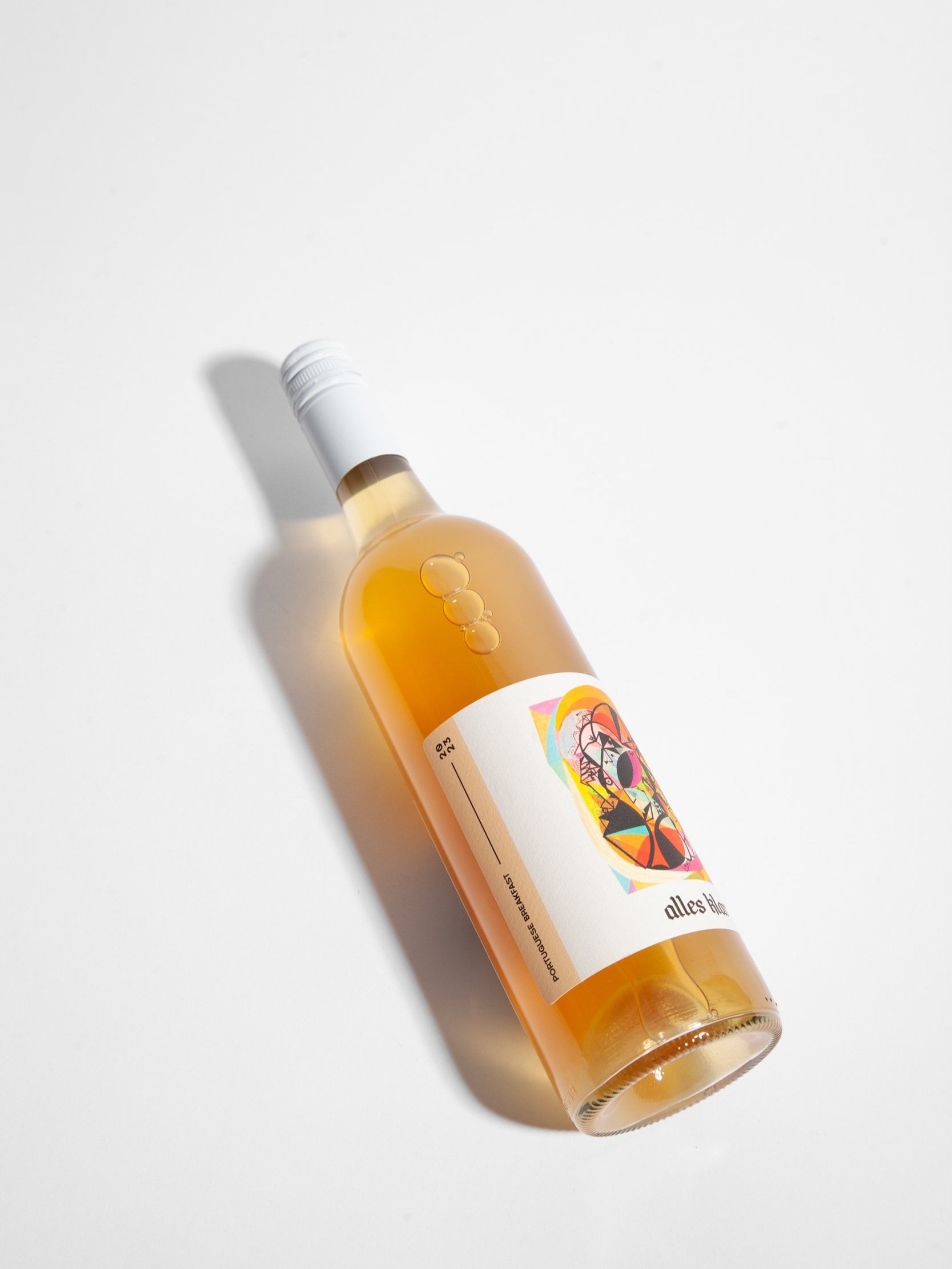 A single bottle of Alles Klar Portuguese Breakfast 2023 Riverland Arinto, a lightly textured crisp white, is shown lying diagonally on a white surface, casting a soft shadow to its left.
