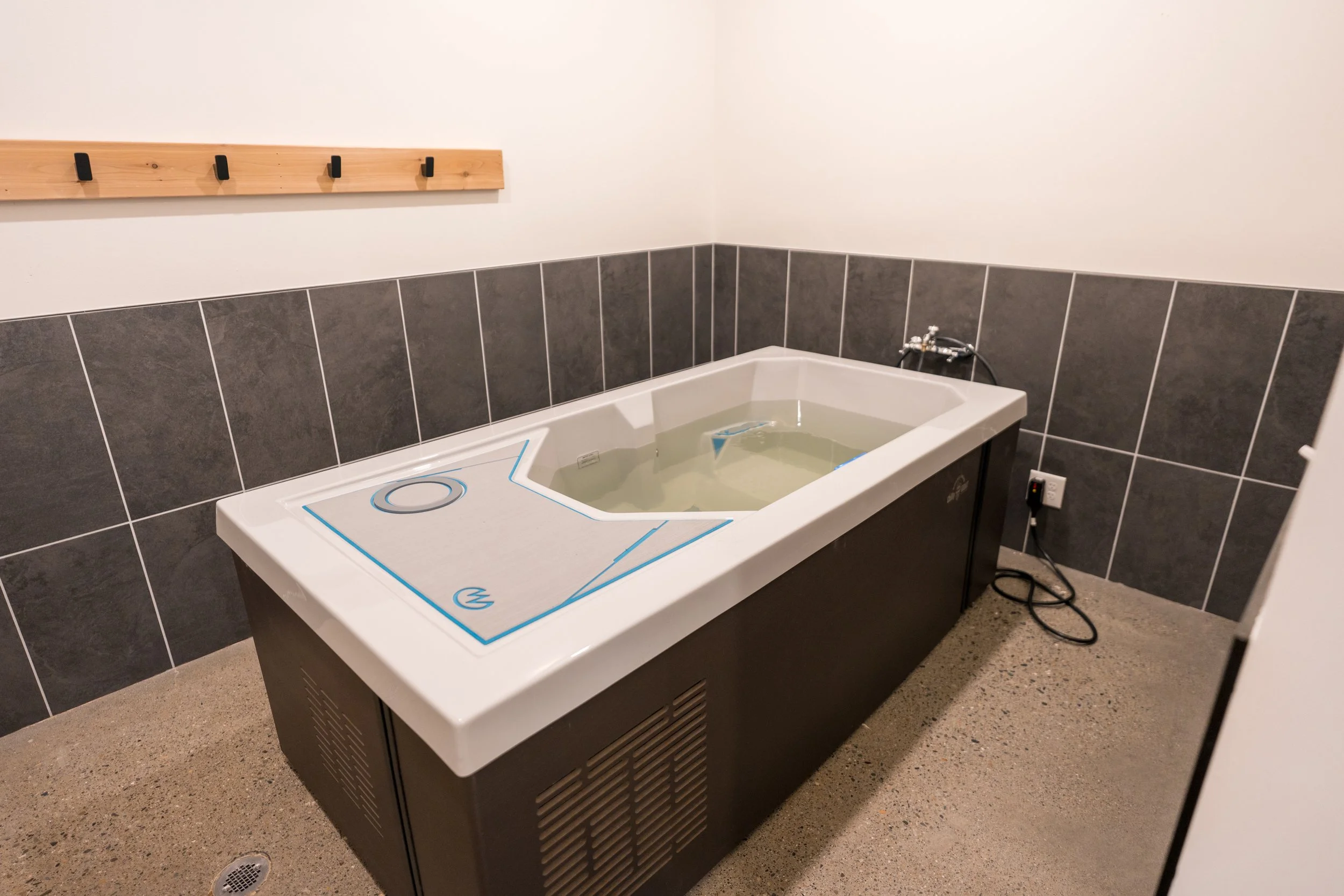 A white, rectangular portable foot spa or whirlpool bath with water, placed on a brown speckled floor. The spa has a control panel with buttons and a circular opening, covered with a plastic film or protective cover. It is positioned against a wall with dark gray tiles, a wooden coat rack with black hooks, and an electrical outlet with a plug.