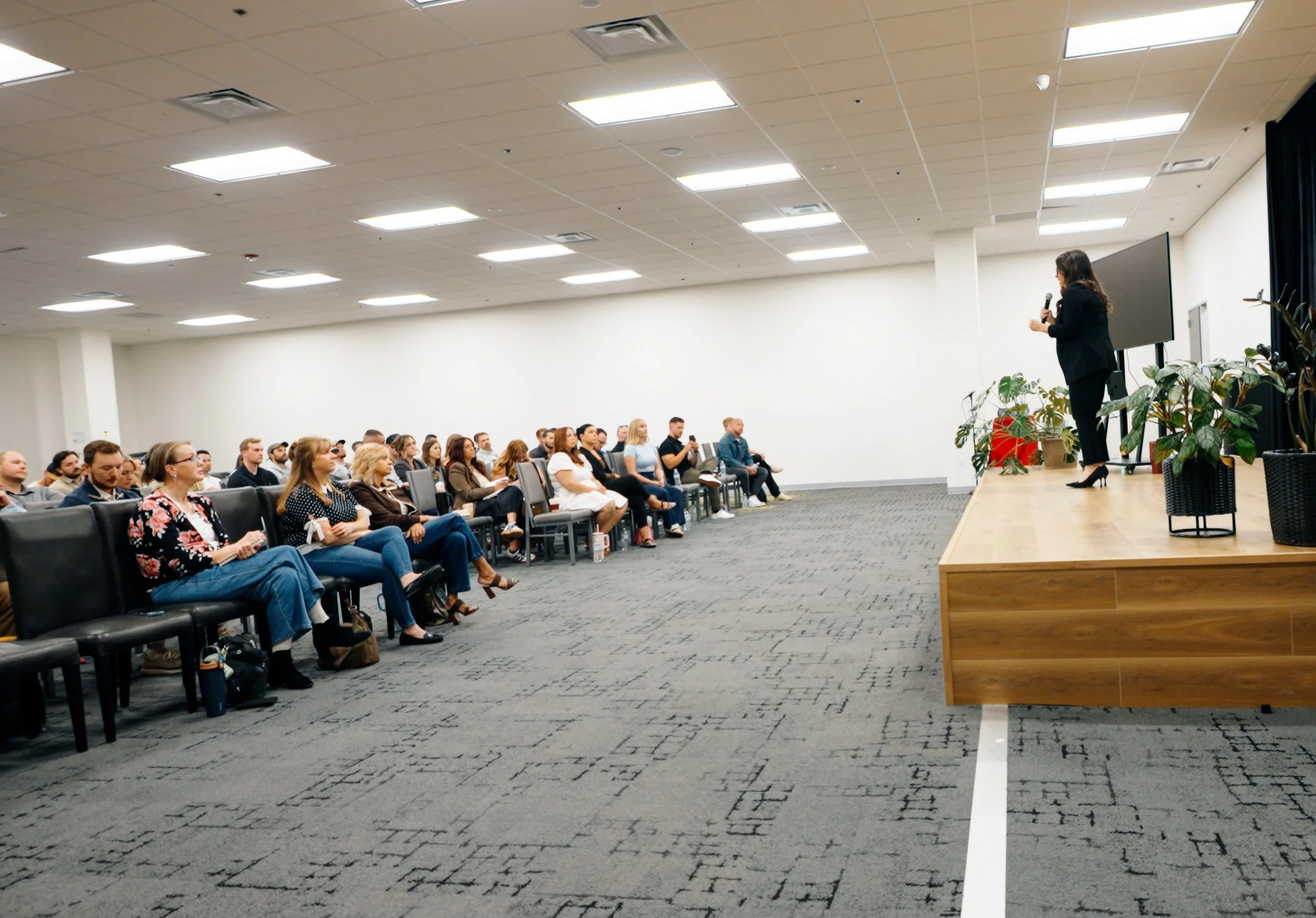A woman in a black suit standing on a stage, speaking into a microphone for an audience seated on chairs in a conference room with a white wall and ceiling lights.