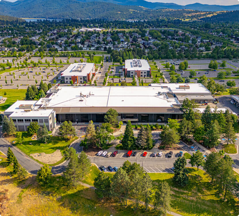 An aerial view of a large modern building with a white roof, surrounded by parking lots, trees, and green lawn areas, with residential neighborhoods and mountains in the distance.